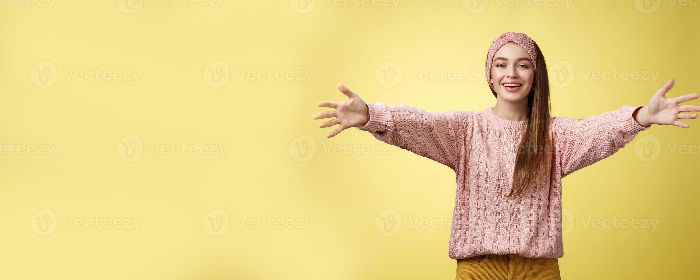 Attractive friendly positive young european woman wearing sweater, headband welcoming extanding arms and smiling at camera giving hugs, embraces friends, cuddling against yellow background photo