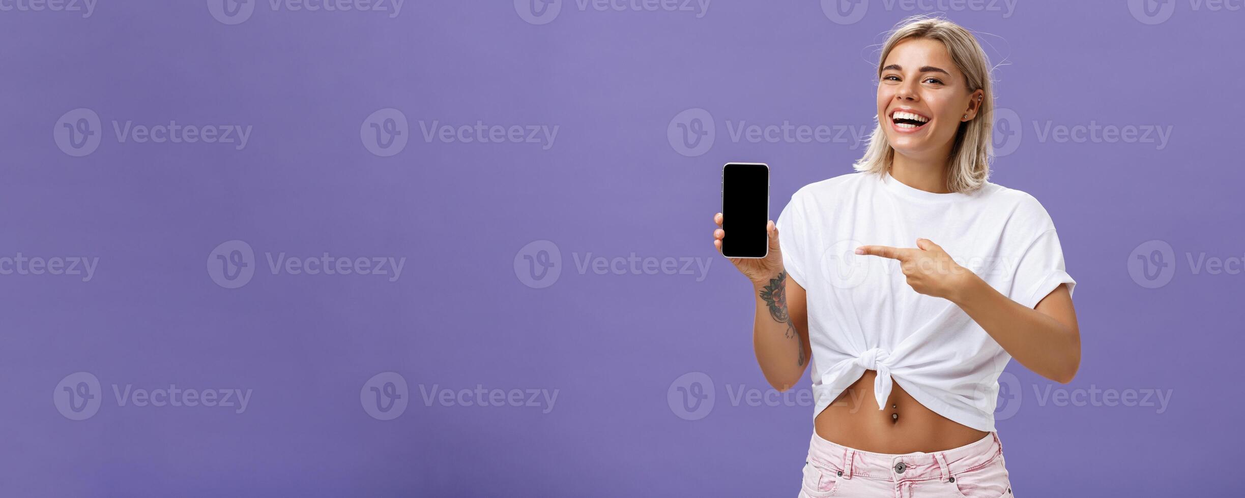 Satisfied happy beautiful tanned woman with blond hairstyle and tattoos holding smartphone showing device screen and pointing at gadget with index finger smiling at camera over purple wall photo