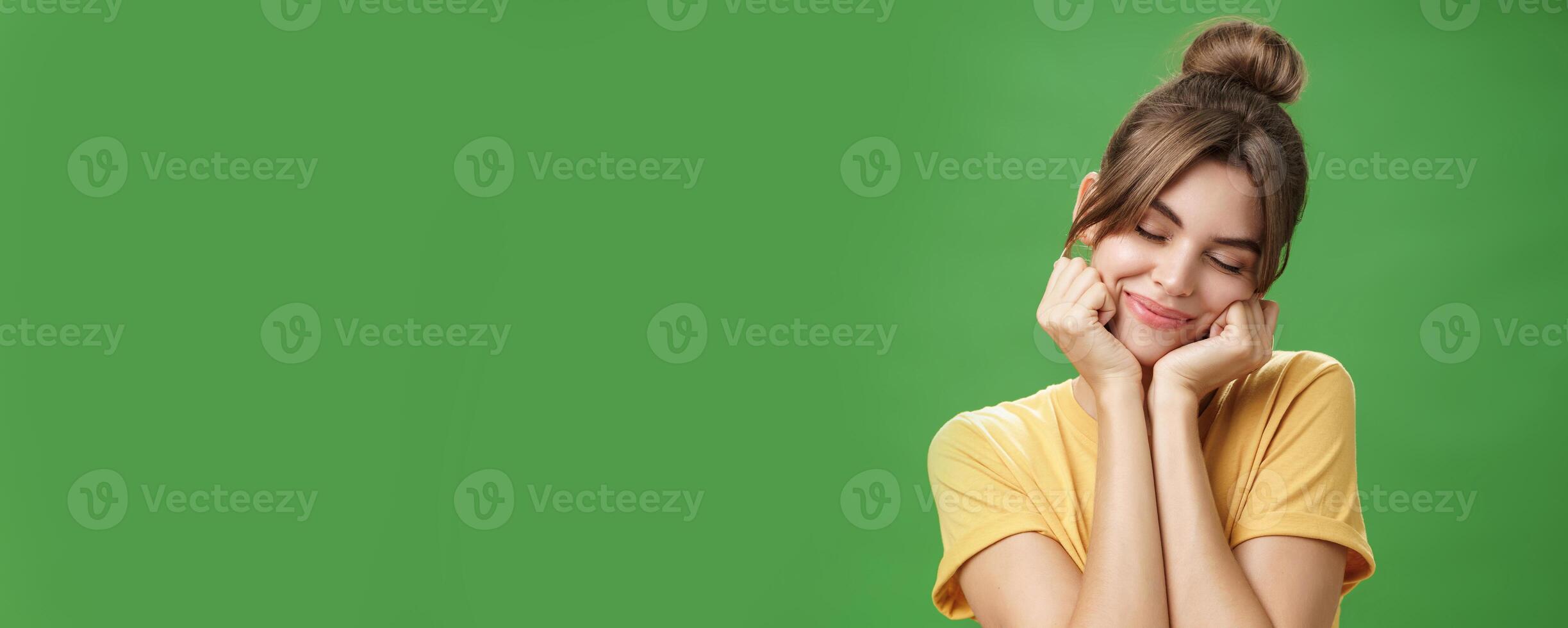 Tender sensual timid european girl in yellow t-shirt leaning head on shoulder touching cheeks with hands closing eyes and smiling with soft grin feeling nostalgic and romantic over green background photo