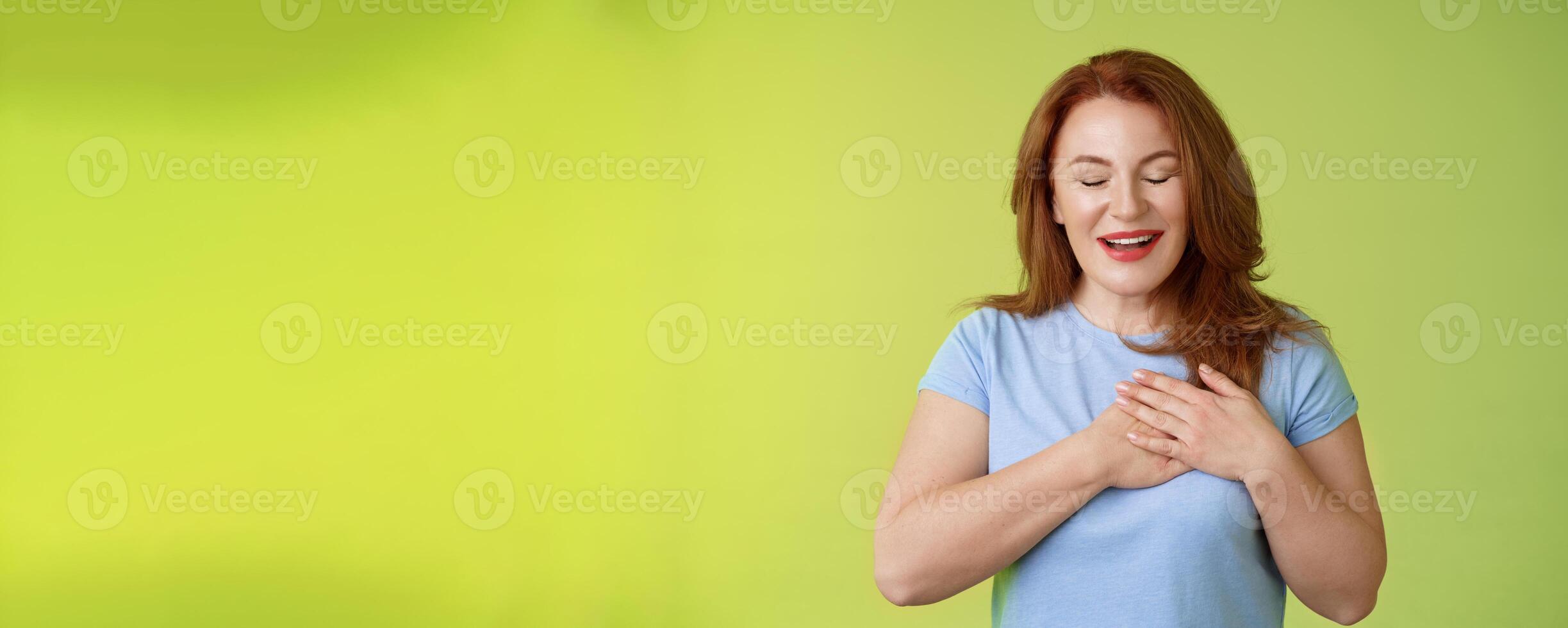 Fascinated cute redhead passionate middle-aged woman sighing lovely touch heart close eyes smiling delighted express admiration temptation feeling appreciation grateful emotions green background photo