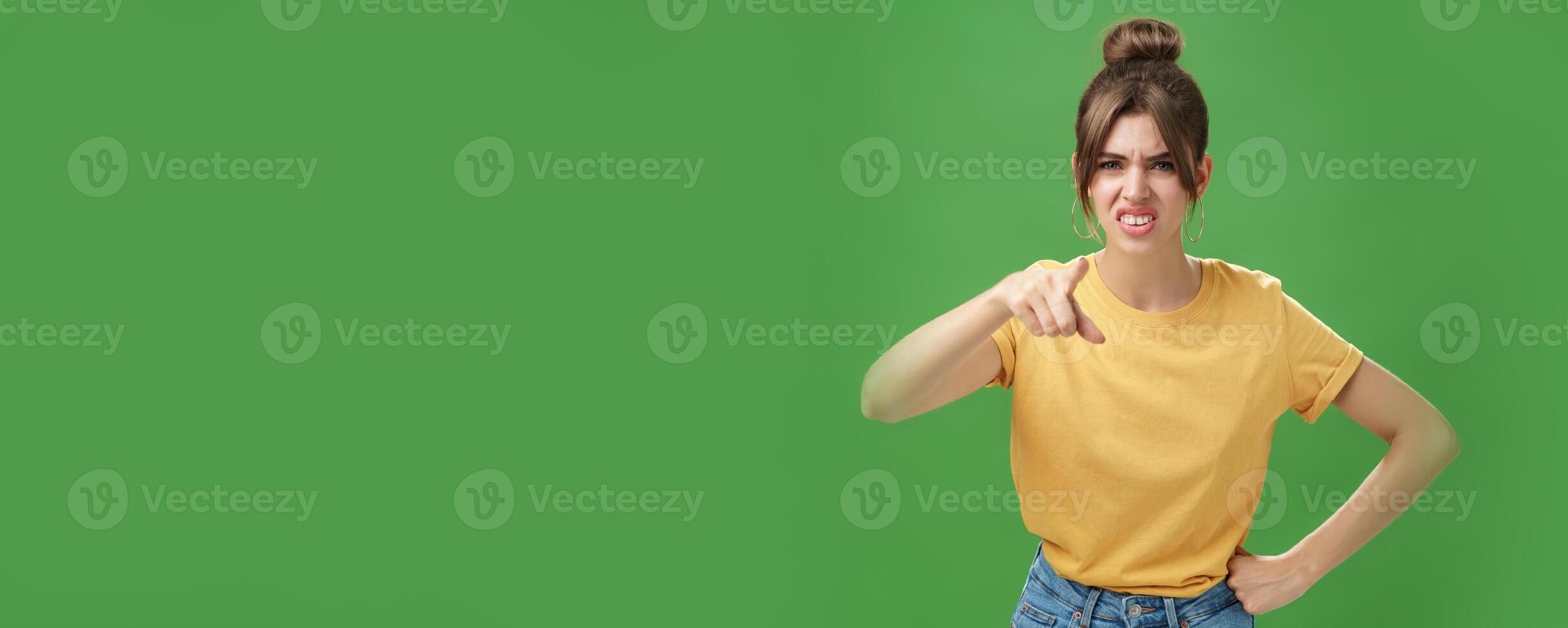 Portrait of arrogant impolite young woman in yellow t-shirt pointing at camera squinting and grimacing from disdain and scorn standing unimpressed judging against green background photo