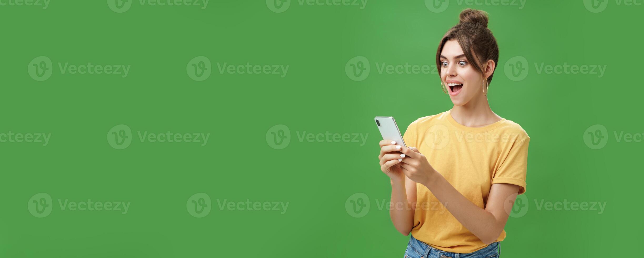 Woman reading surprising satisfying message in smartphone opening mouth from excitement, smiling amazed looking astonished at cellphone screen posing against green background in casual yellow t-shirt photo