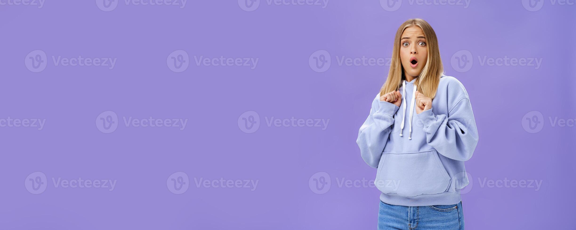 Woman scared of sudden loud noise in dark gasping looking shocked and concerned tilting backwards with frightened look pressing clenched fists to body from fear, wearing warm hoodie over purple wall photo