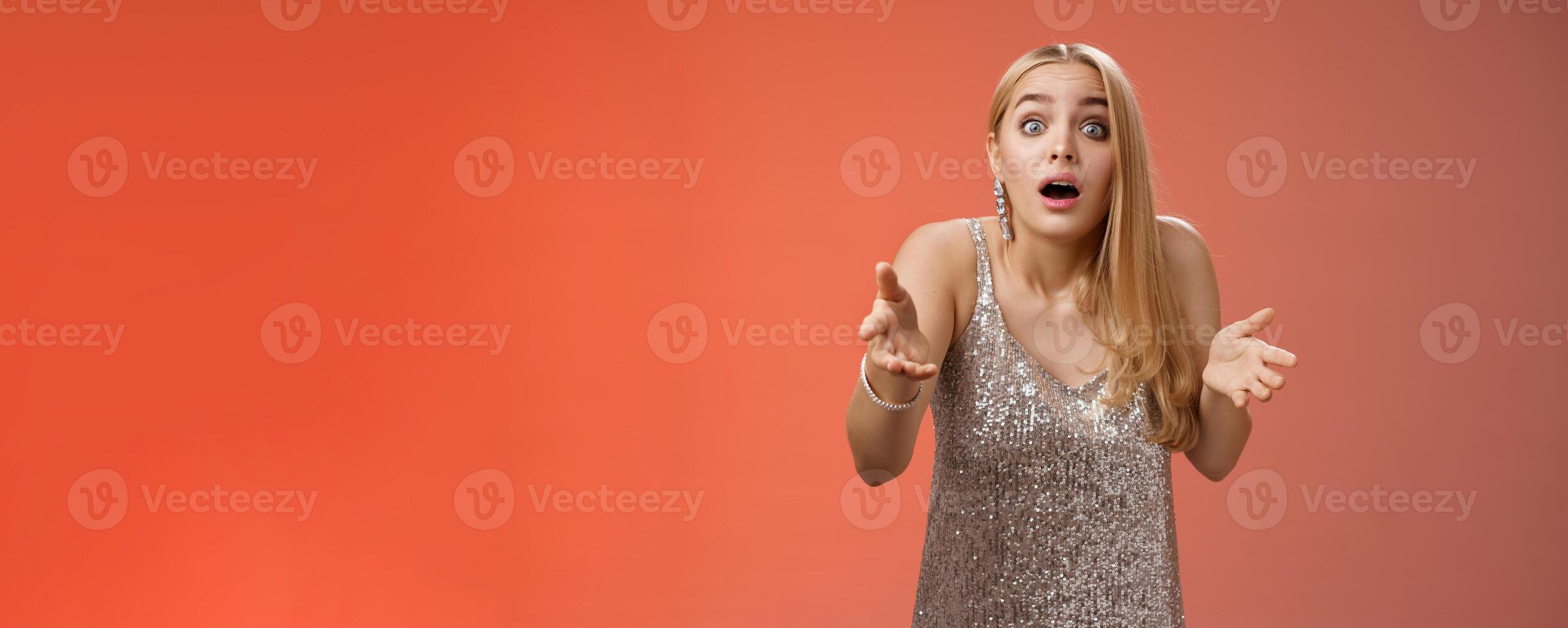 Impressed overwhelmed blond woman retelling exciting story gesturing astonished widen eyes shrugging explaining what happened talking passionately in silver glamour dress standing red background photo