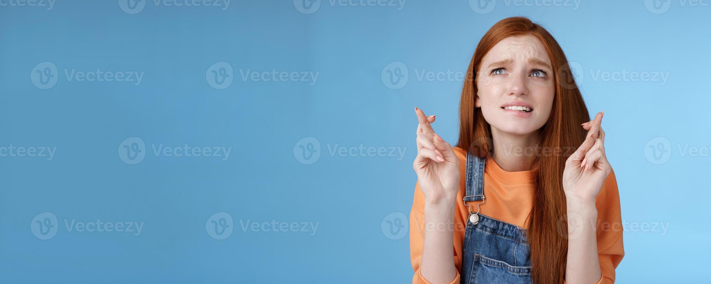 Worried unconfident young redhead intense cute girl frowning nervously waiting improtant moment cross fingers good luck bite lip anxiously frowning look upper left corner praying wish come true photo