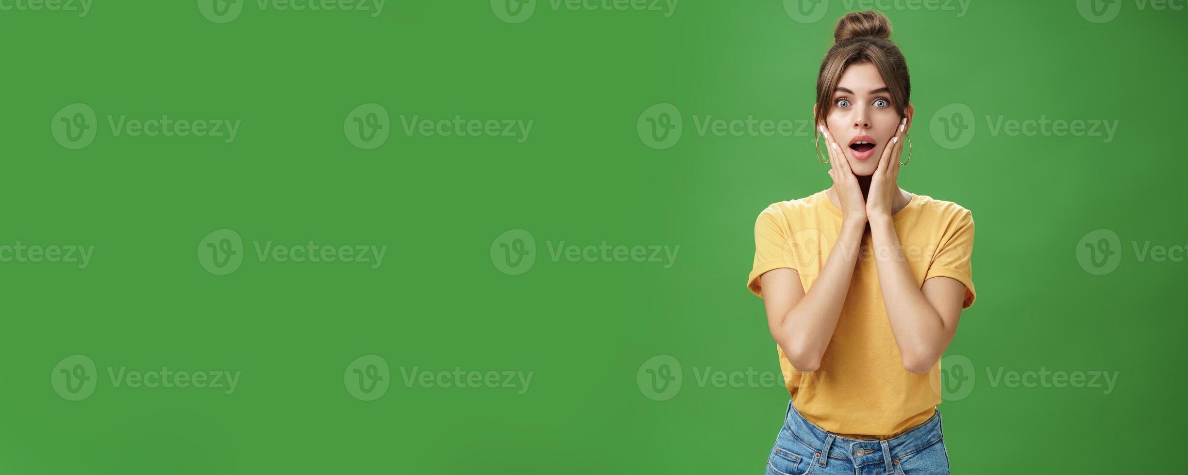 Woman feeling surprised and delighted enjoying good result after detox and applying facial skincare mask touching cheeks opening mouth from amazement and excited posing over green background photo