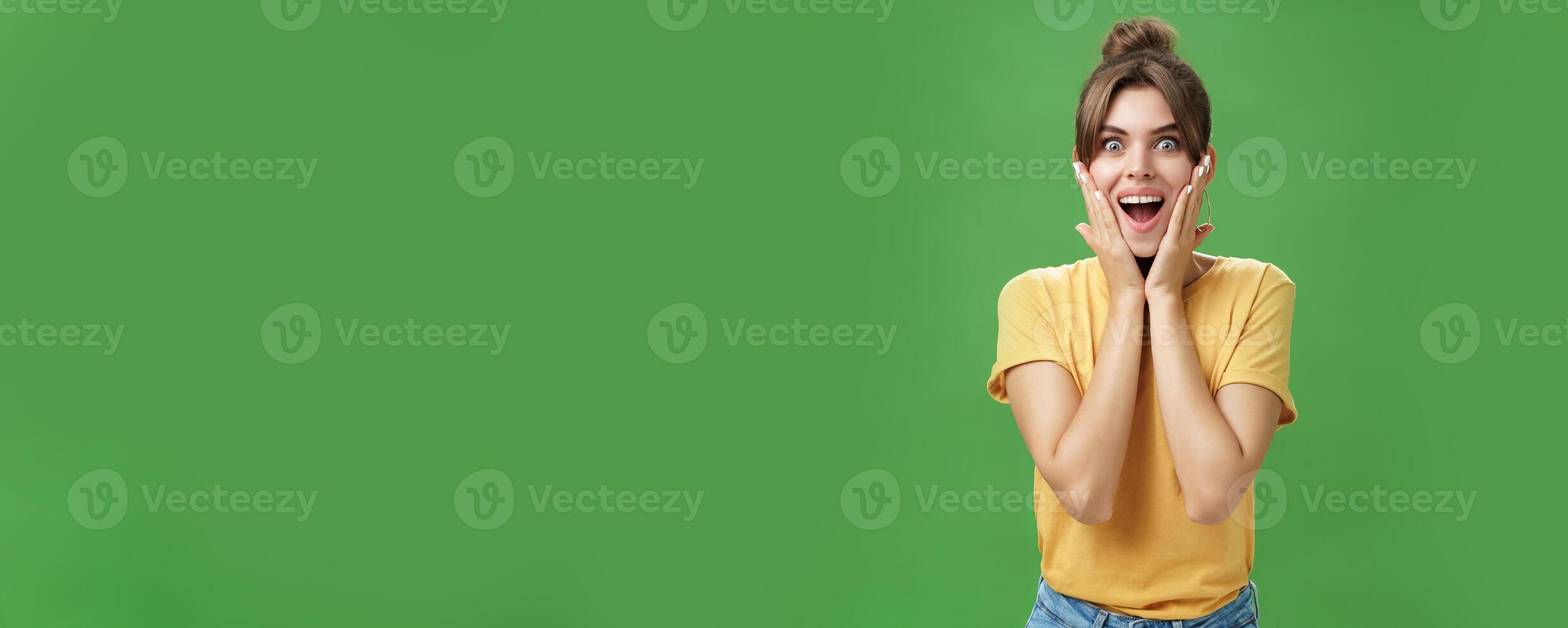Indoor shot of excited and surprised energized attractive glamorous female in yellow t-shirt touching cheeks from amazement smiling broadly astonished posing over green background photo