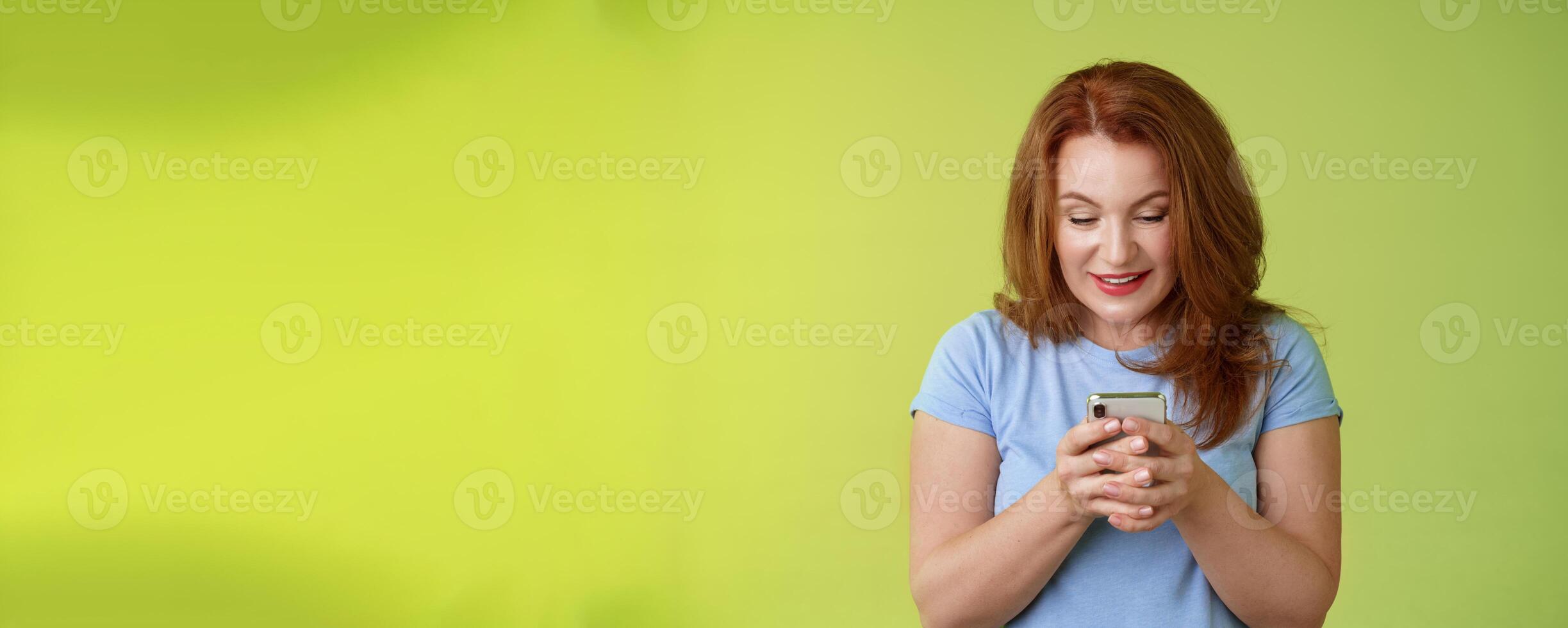 Intrigued excited tempting redhead middle-aged woman shopping online smartphone hold mobile phone look pleased entertained gadget screen smiling delighted playing game found perfect app photo