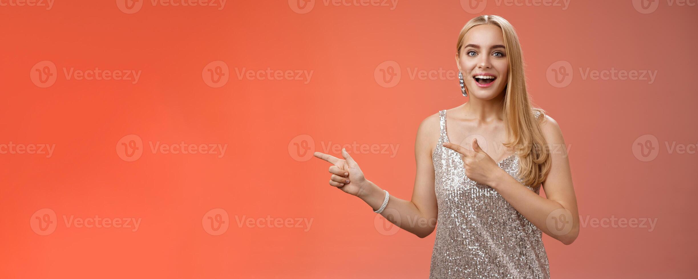 Excited carefree happy young attractive woman joyful attend awesome party in silver beautiful glittering dress pointing left suggesting come together inviting dance smiling cute red background photo