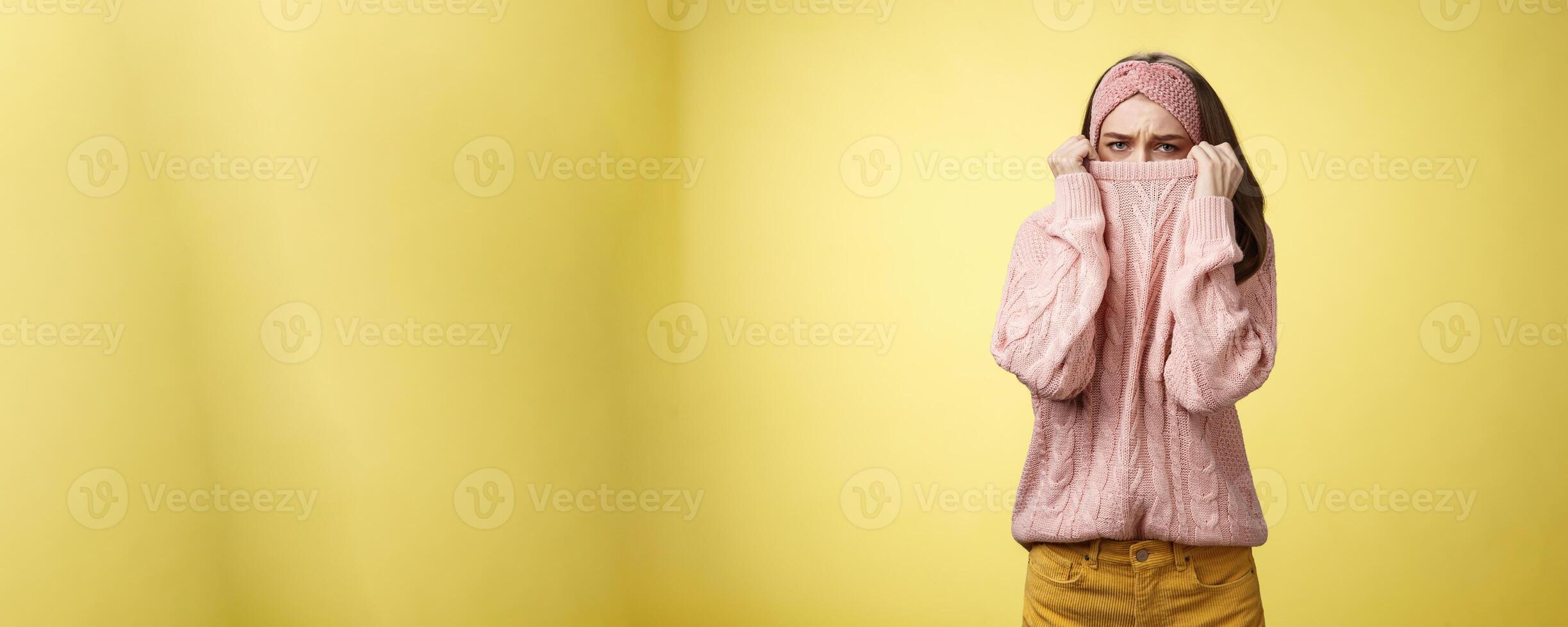 Sad silly, gloomy cute young girl hiding face in sweater collar pulling clothes on nose frowning displeased, unhappy, looking indignant and reluctant standing disappointed over yellow background photo