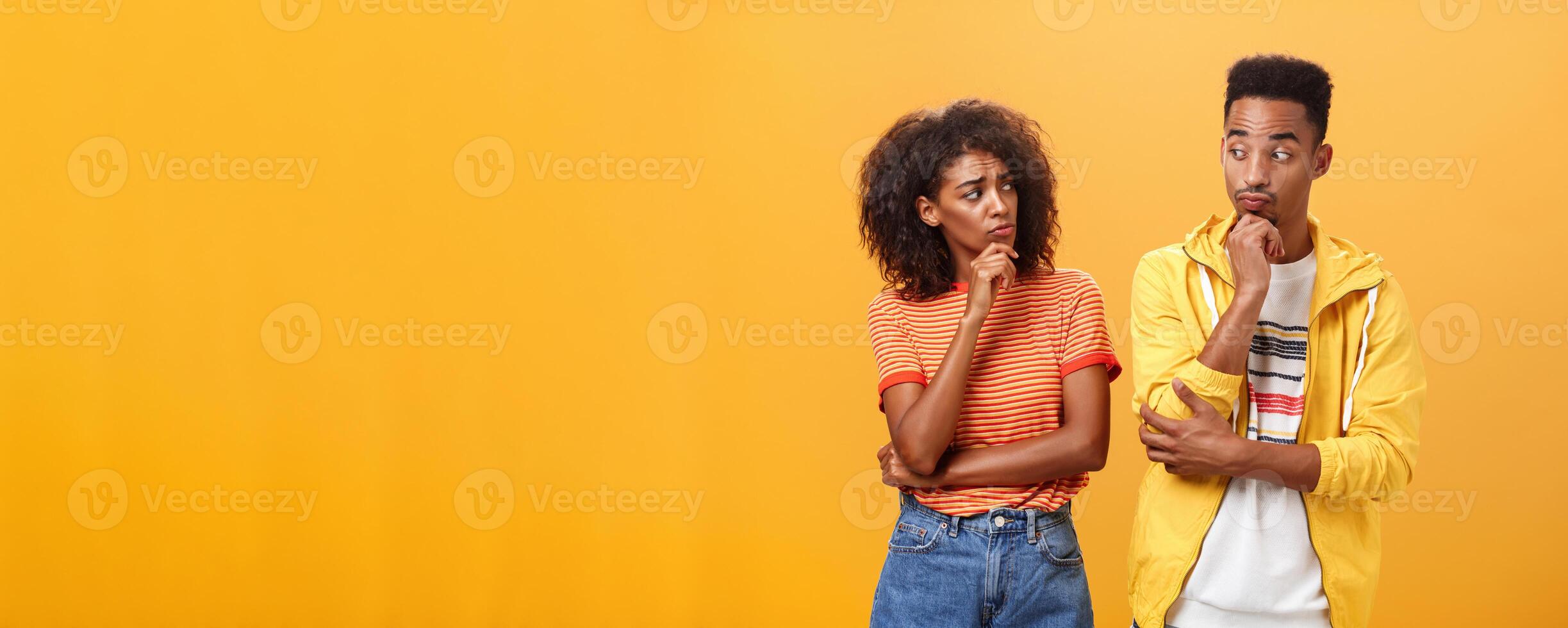Silly confused girl standing gloomy and worried pursing lips and frowning holding hand on chin looking at boyfriend confused while guy glancing at girlfriend with curious expression over orange wall photo