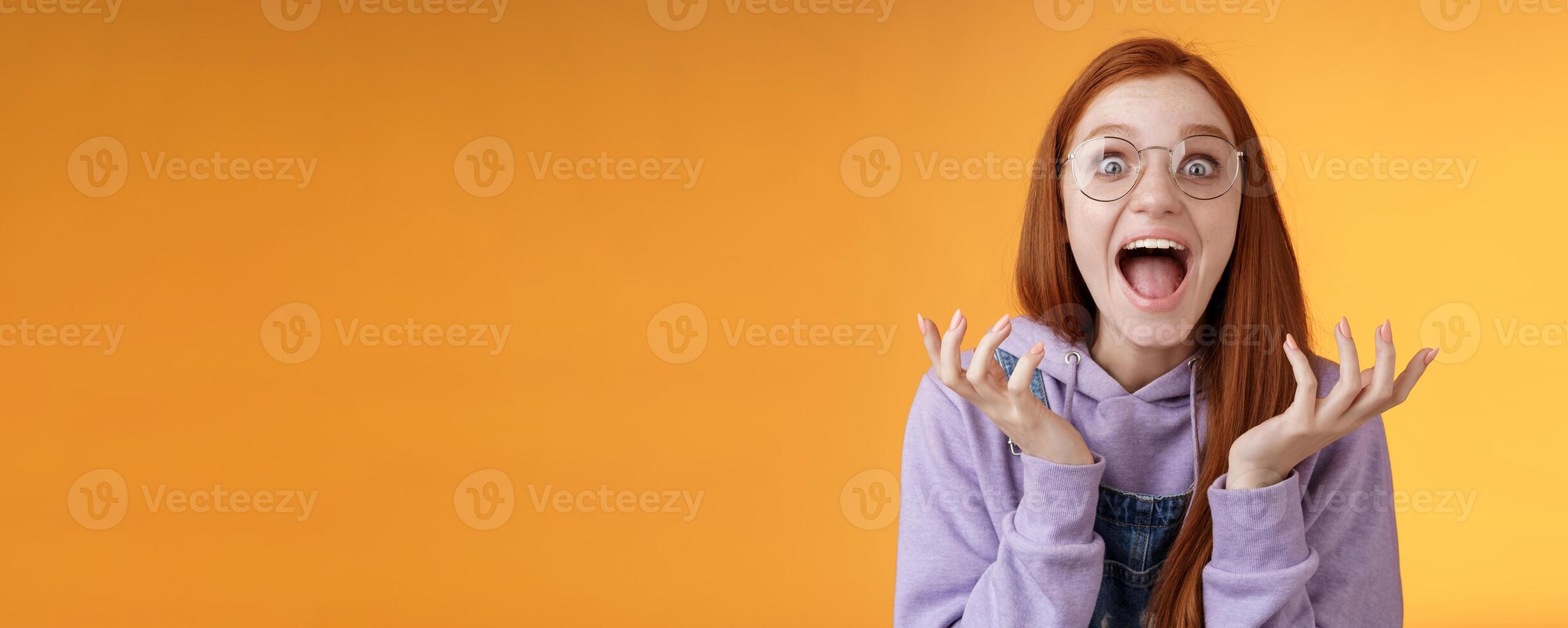 Shocked excited overwhelmed young screaming happy redhead girl wearing glasses winning hear excellent news yelling out loud rejoicing unbelievable luck raising hands spread stare surprised photo