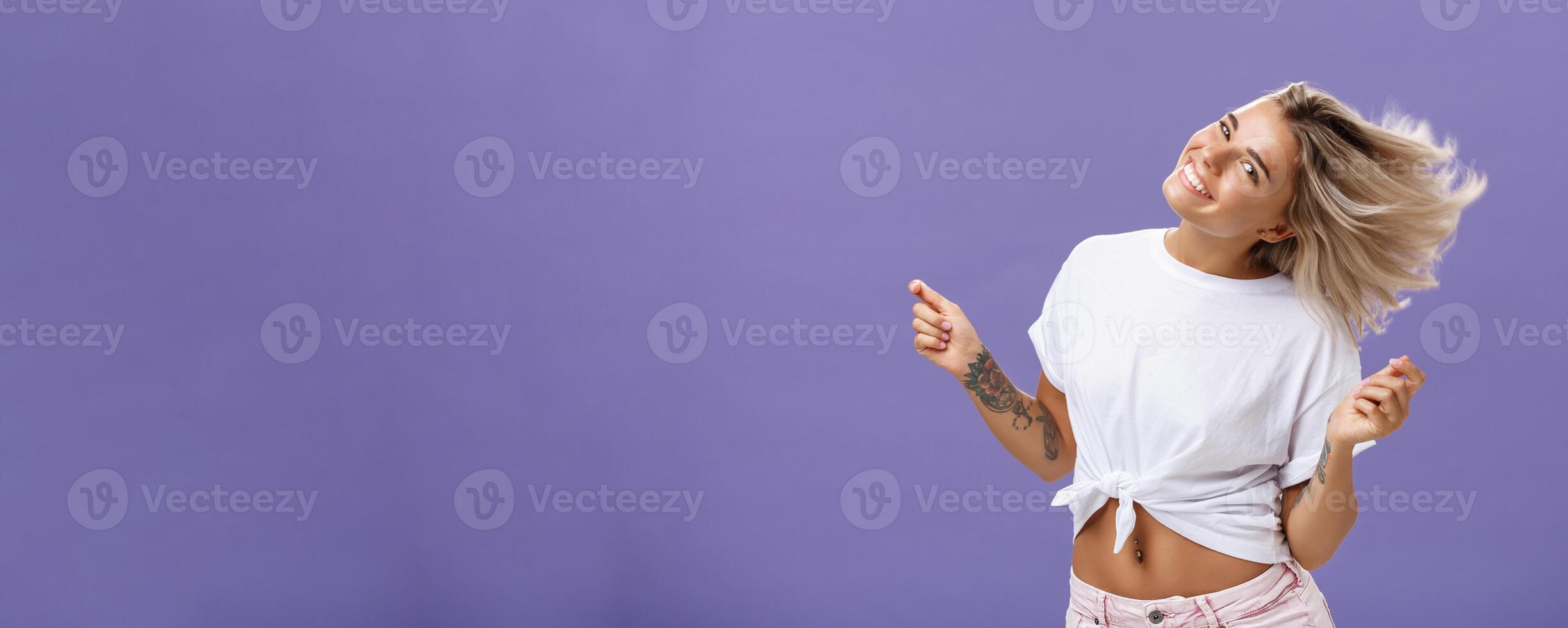 Portrait of happy carefree attractive woman dancing and waving hair from rejoice and cheer smiling broadly tilting head celebrating end or working day enjoying nice sunny day on beach over purple wall photo