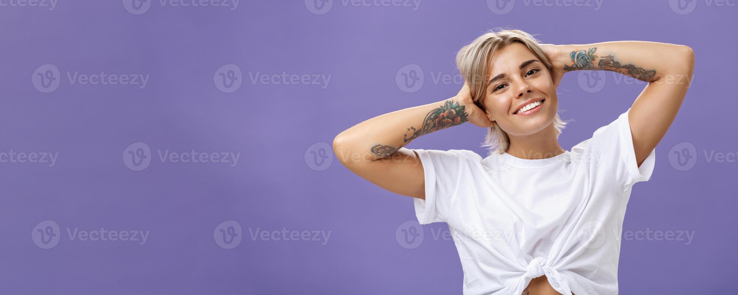 Close-up shot of good-looking stylish and relaxed blond woman with tattoos on arms holding hands behind head smiling with carefree pleased look enjoying weekends over purple background photo