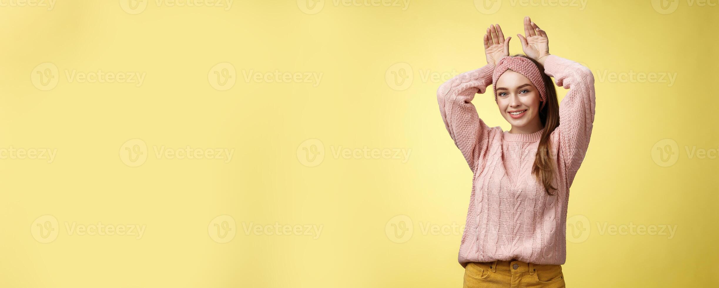Nice charming playful young european female having fun, playing mimicking bunny showing rabbit ears with hands behind head, smiling cute, tender, happy greeting boyfriend welcome back ove yellow wall photo