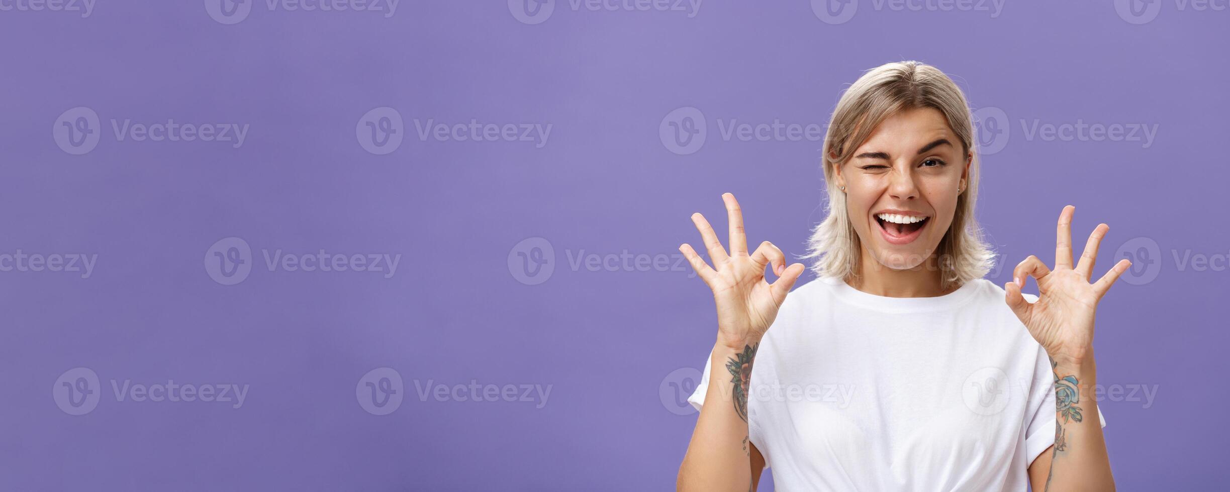 Cute attractive online store owner assuring customers delivery come in time. Delighted good-looking young female with tanned skin and tattoos showing okay or excellent gesture and winking with joy photo