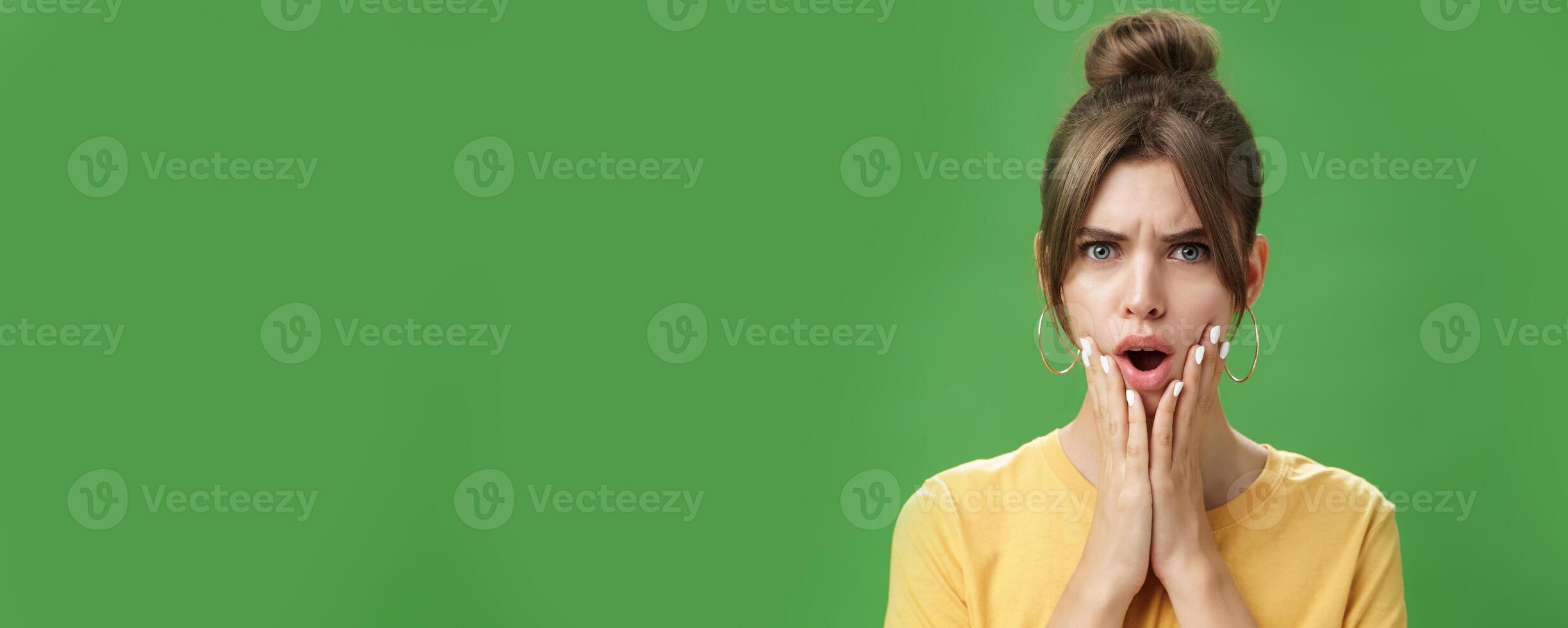 Shocked woman reacting to pimple looking disturbed and displeased in mirror holding hands pressed to cheeks open mouth and frowning being disappointed and upset over green background photo
