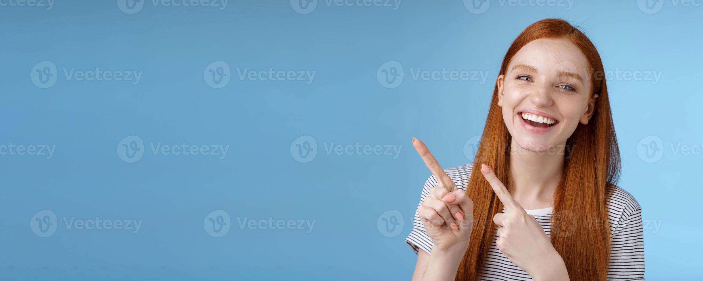 Happy carefree smiling redhead caucasian girl ginger straight hairstyle pointing upper left corner laughing joyfully amused cool promo giving recommendation try new product, standing blue background photo