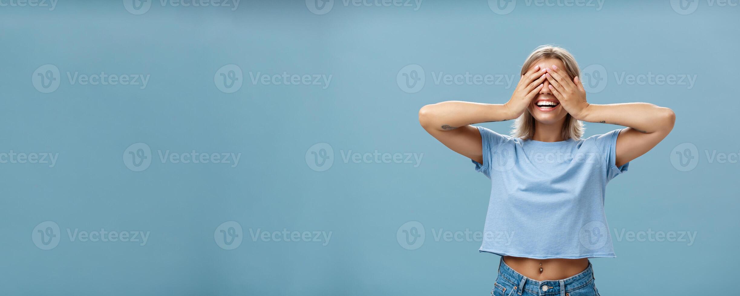 Hide if you can. Portrait of carefree and happy good-looking playful girl with blonde hair smiling broadly while counting ten and playing hide-and-seek with closed eyes over palms near blue wall photo