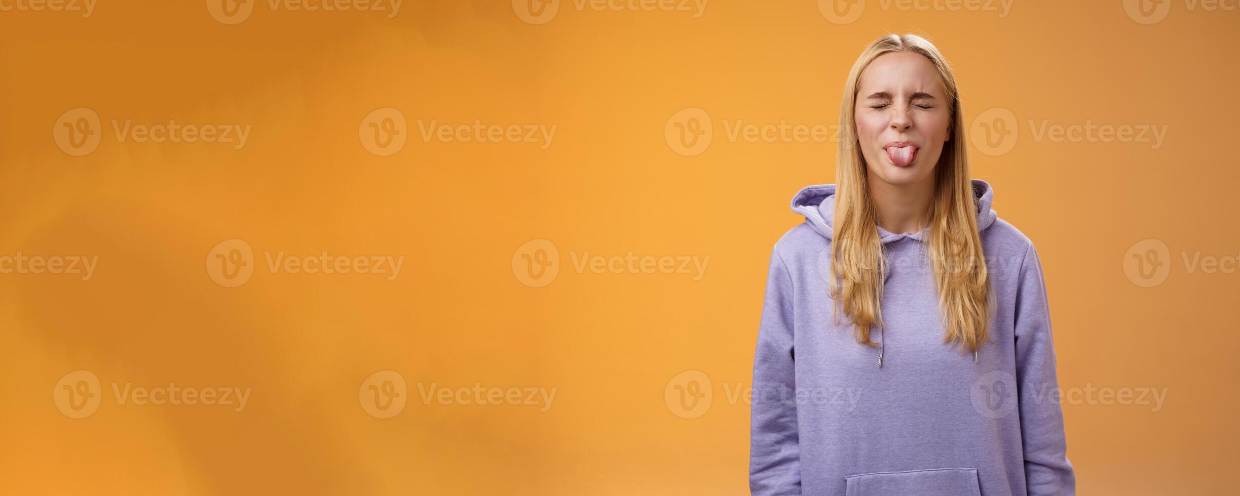 Playful amused charismatic young funny blond girl having fun close eyes showing tongue rebellious fool around unbothered enjoying carefree sunny spring days standing orange background photo