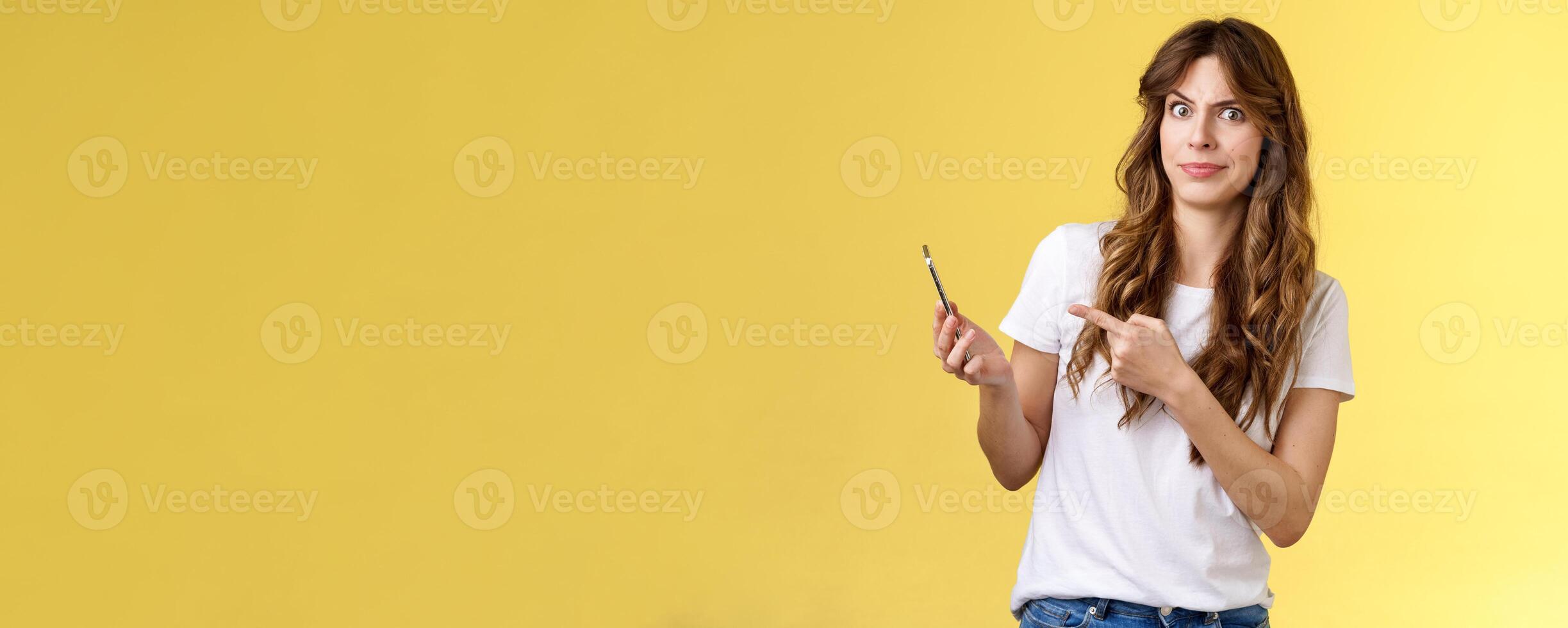 Girl answers strange weird call receive crazy upsetting message cringe doubtful displeased smirking dismay pointing suspicious smartphone stand yellow background intense frustrated photo