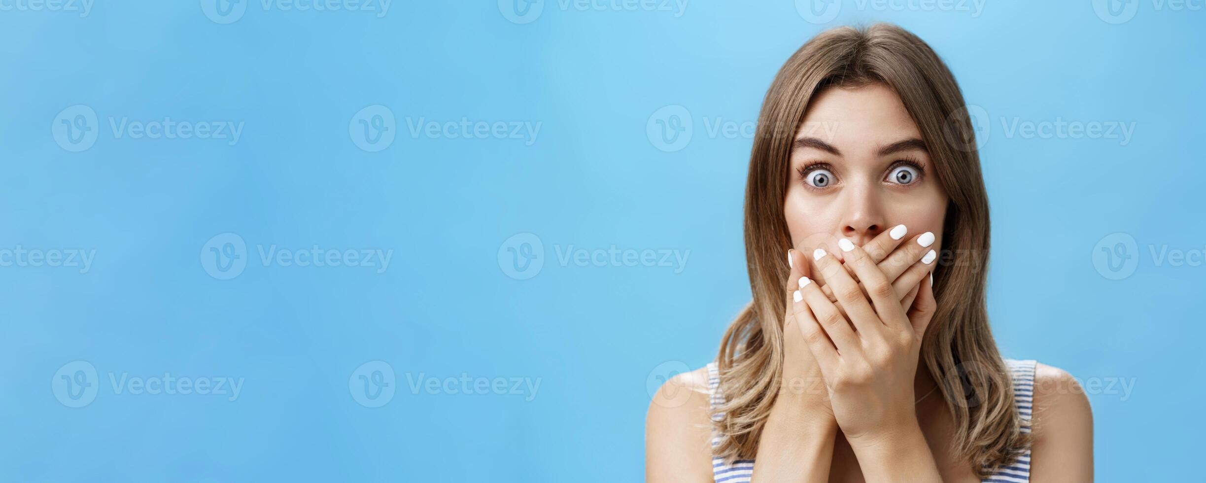 Close-up shot of shocked speechless woman covering mouth with pressed palms popping eyes at camera from surprise and worry, gasping witnessing terrible scene, feeling scared and intense over blue wall photo
