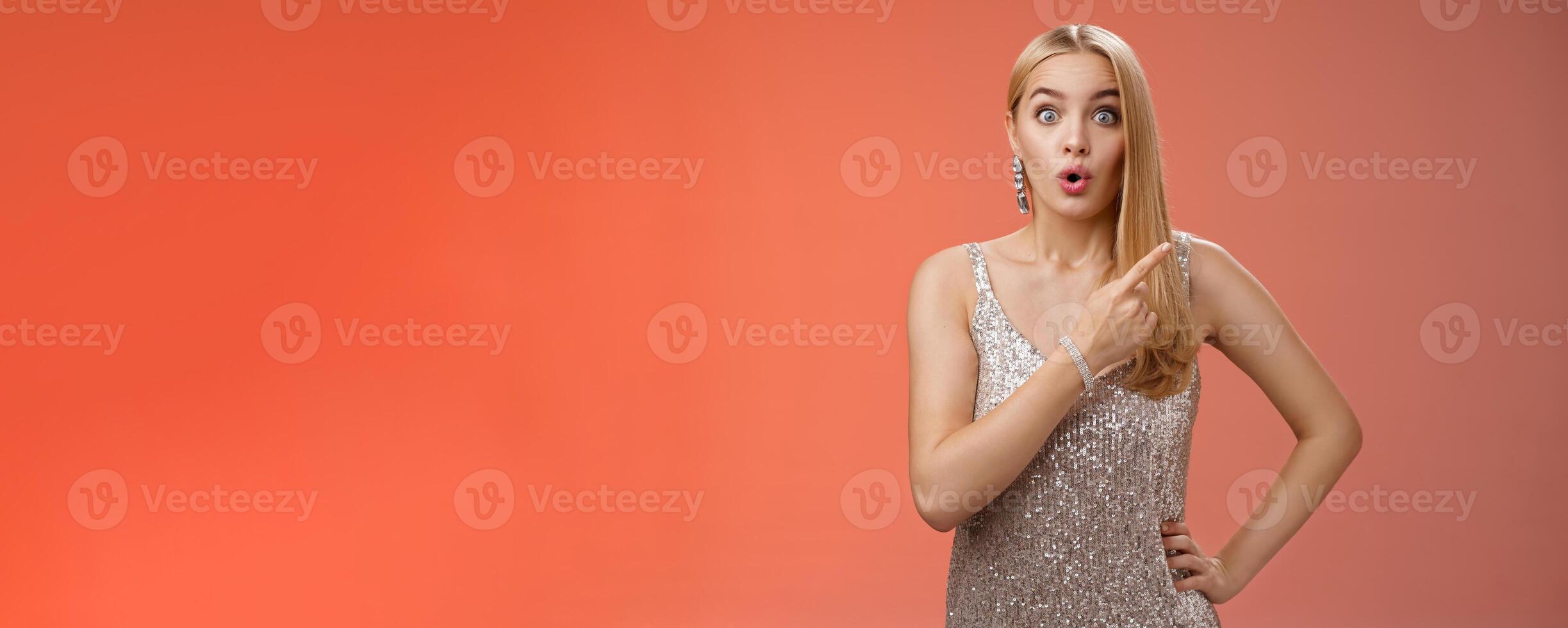 Wondered amused attractive blond woman talking party widen eyes say wow pointing questioned right direction see famous person standing surprised impressed gaze camera, red background photo