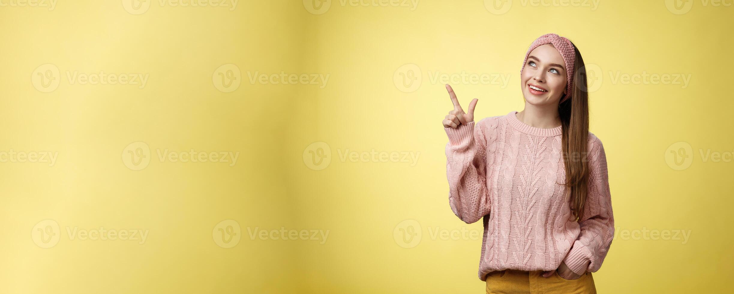 Portrait of amused interested cute 20s european girl wearing sweater, headband looking upper left corner pointing sideways smiling intrigued, ecstatic, captured by curious promo over yellow wall photo