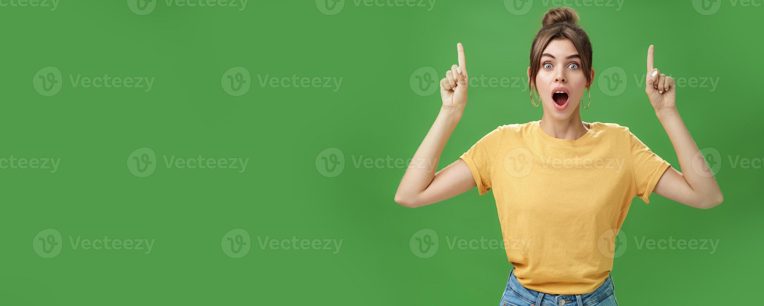 Waist-up shot of interested and surprised amazed good-looking female in yellow t-shirt with tattoo dropping jaw impressed and astonished raising hands pointing up questioned against green background photo