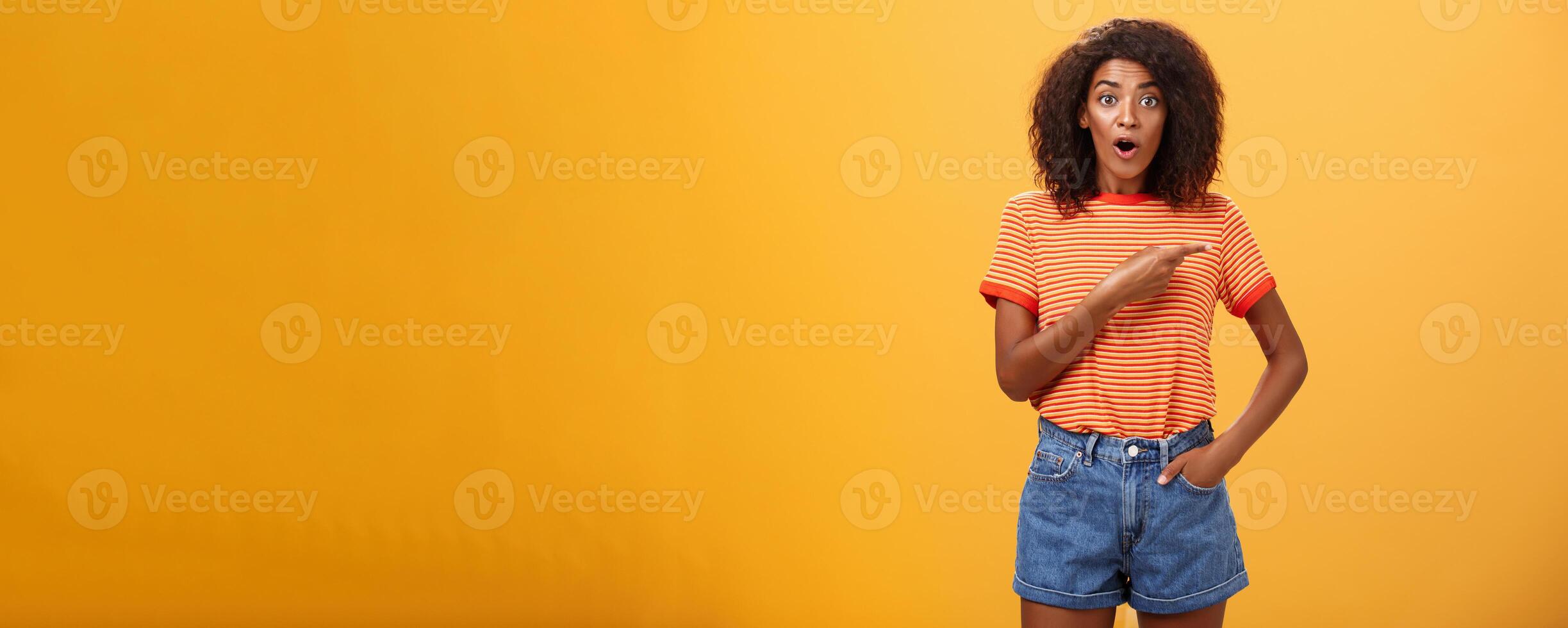 Studio shot of stylish fit female model in denim shorts and t-shirt gasping impressed pointing right while asking question being interested during observing masterclass over orange background photo