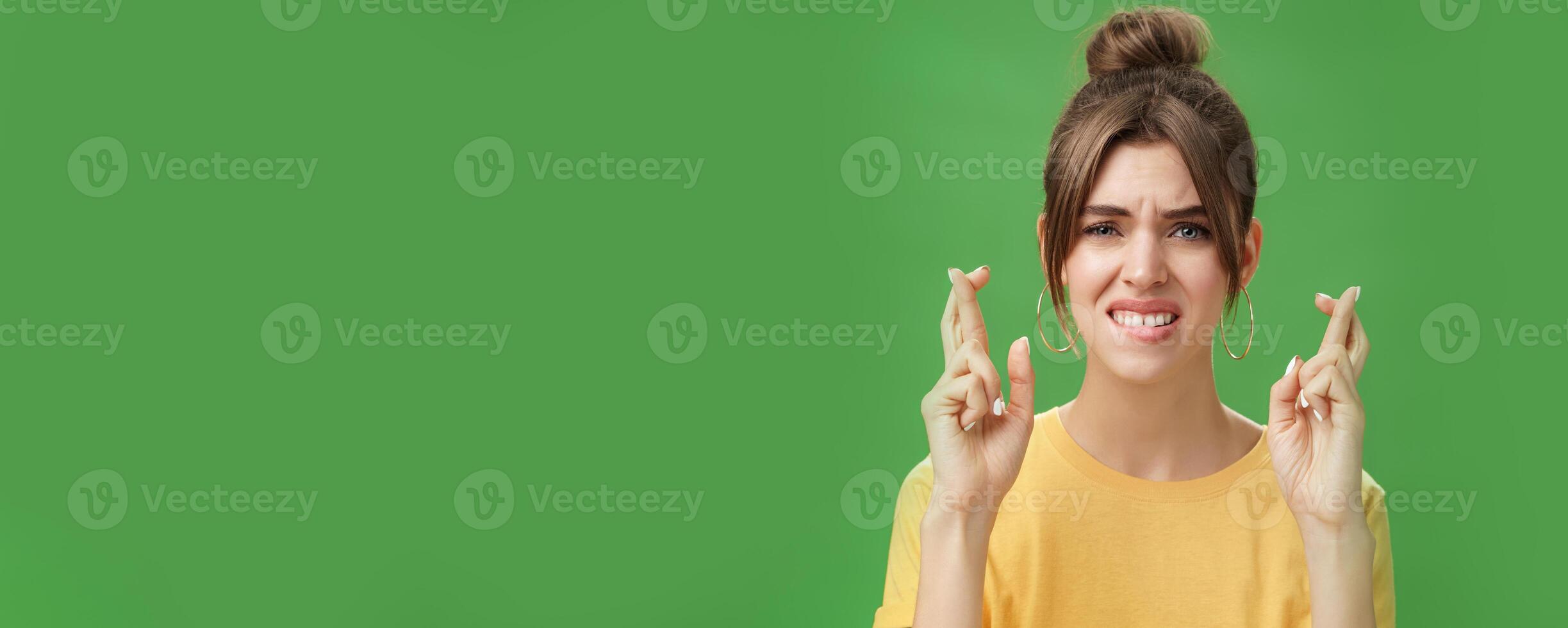 Close-up shot of nervous woman praying for having positive answer crossing fingers for good luck biting lower lip anxiously and frowning being worried while hoping to get job over green background photo