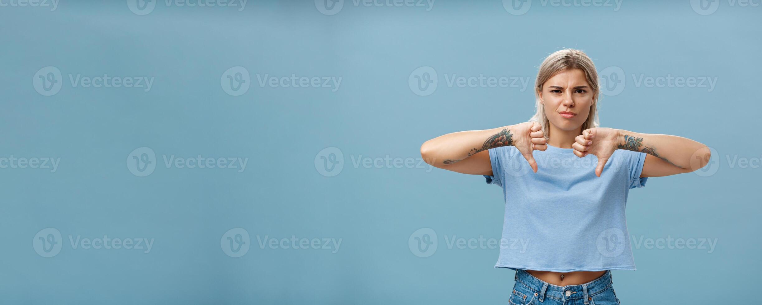 No way mate dislike. Portrait of dissatisfied bossy female tattoo artist with tattoos on arms frowning from displeasure showing thumbs down in disapproval standing over blue background photo