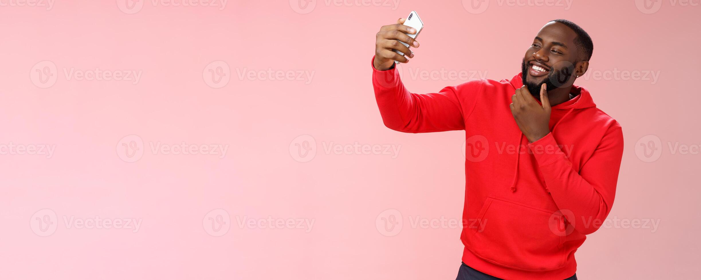 Cheeky cool african american confident bearded guy in red hoodie taking selfie update date app profile photo holding smartphone raised touching face sassy smiling look phone display flirty