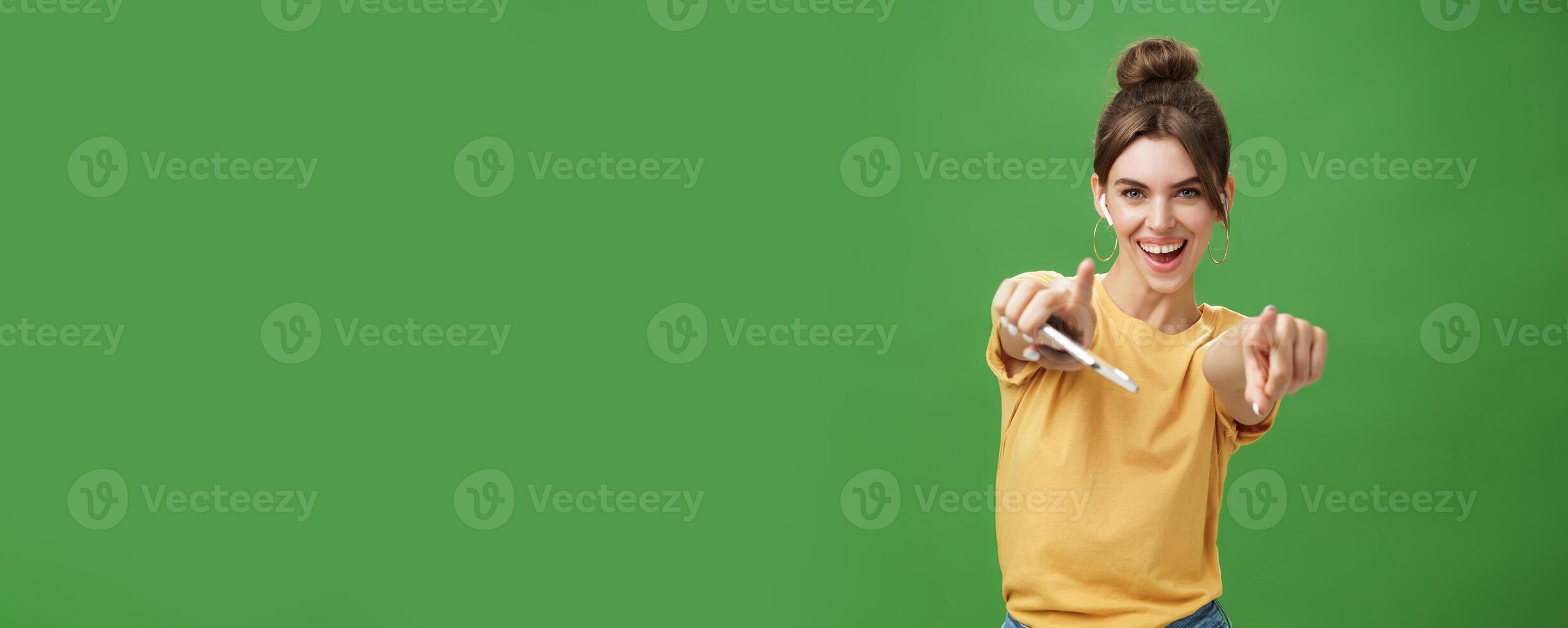 Energized charismatic attractive young woman with combed hair and cute diastema holding smartphone dancing joyfully listening music in wireless earphones pointing and smiling at camera over green wall photo