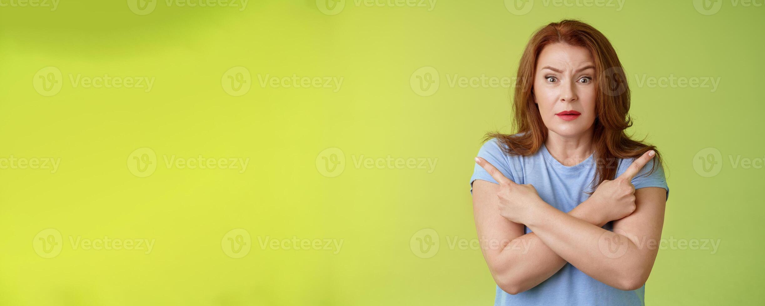 Help me choose. Worried unsure doubtful cute redhead middle-aged silly woman asking advice cross arms body pointing sideways left right look concerned camera make difficult decision green background photo