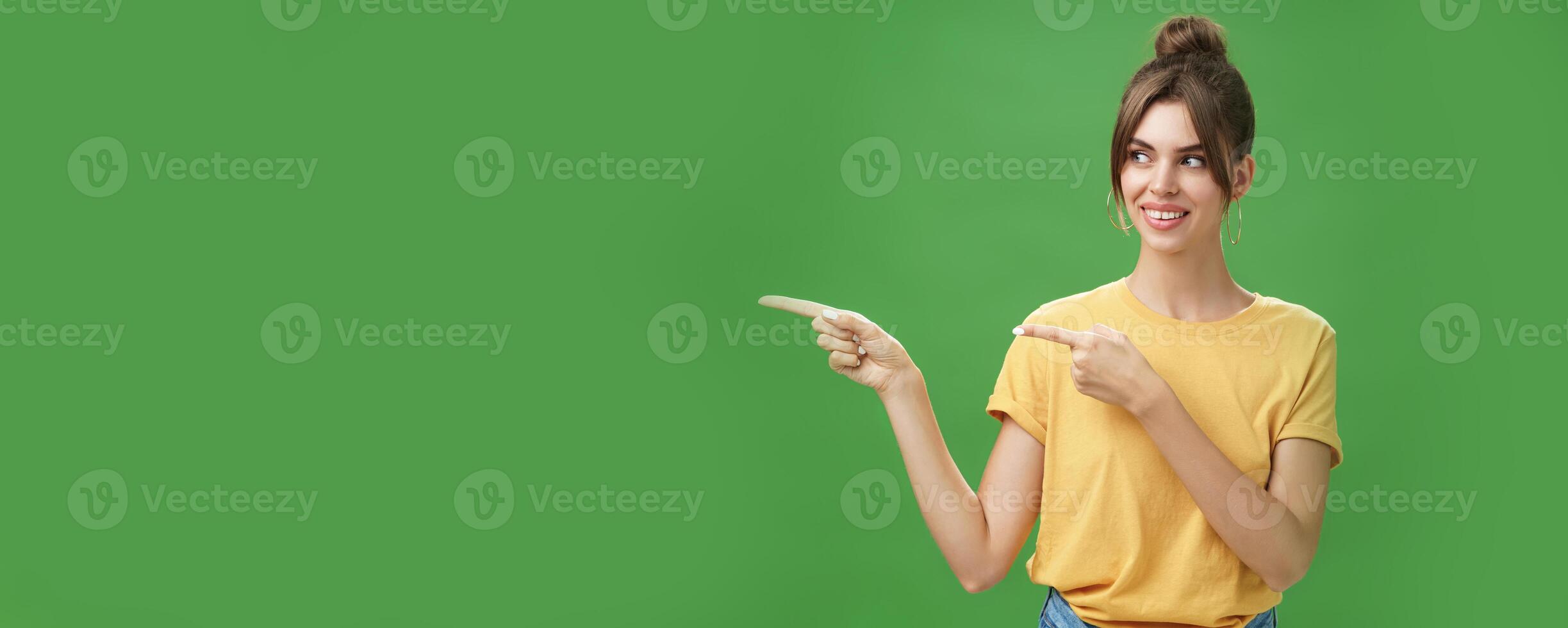 Waist-up shot of good-looking friendly and feminine female coworker in yellow t-shirt showing newbie office pointing and looking left with cute happy smile posing against green background photo