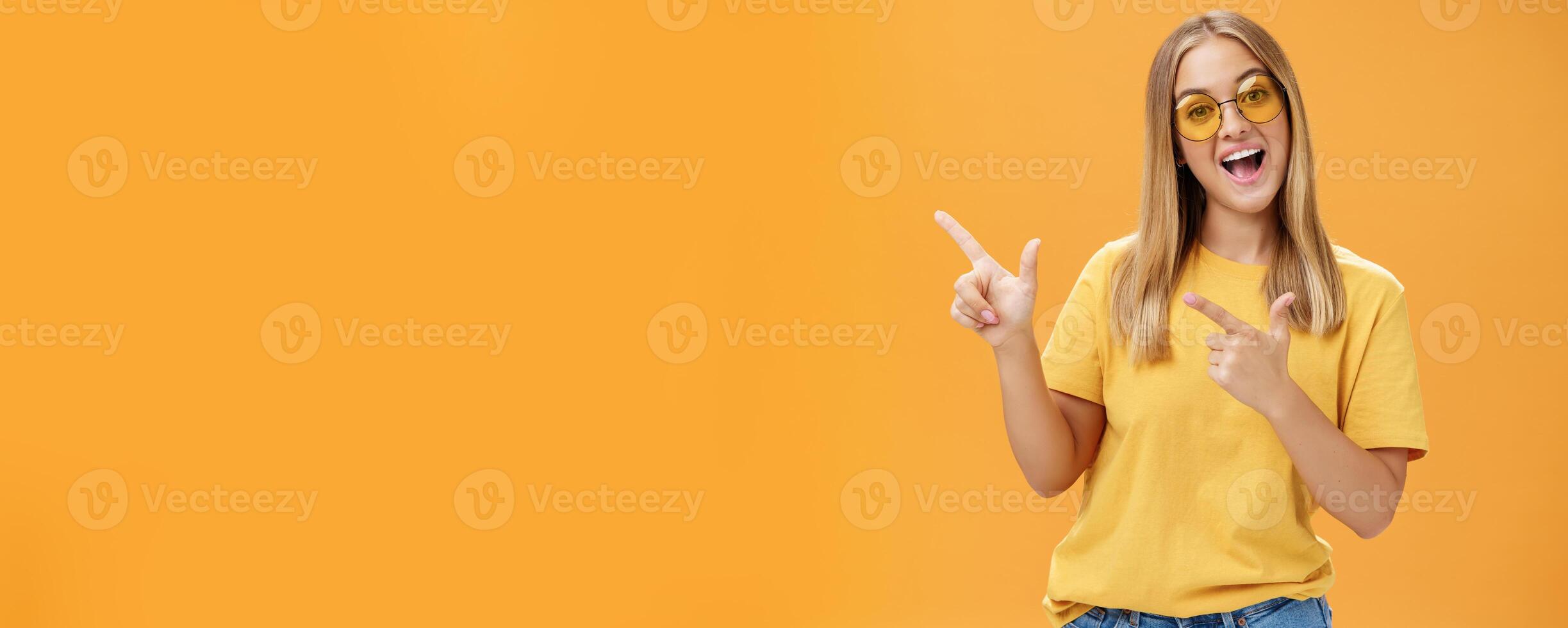 Click her watch my videos. Happy confident and outgoing charming young girl with straight hair in sunglasses and yellow t-shirt pointing at upper left corner promoting product with joyful smile photo