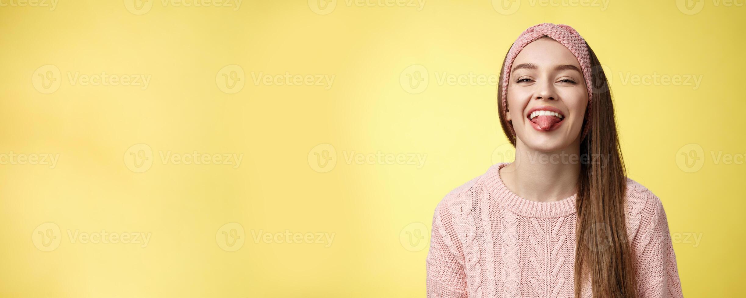 Positive entertained cute funny glamour young european girl in sweater, knitted trendy headband smiling fooling around showing tongue playfully, mocking friend enjoying sunny day over yellow wall photo