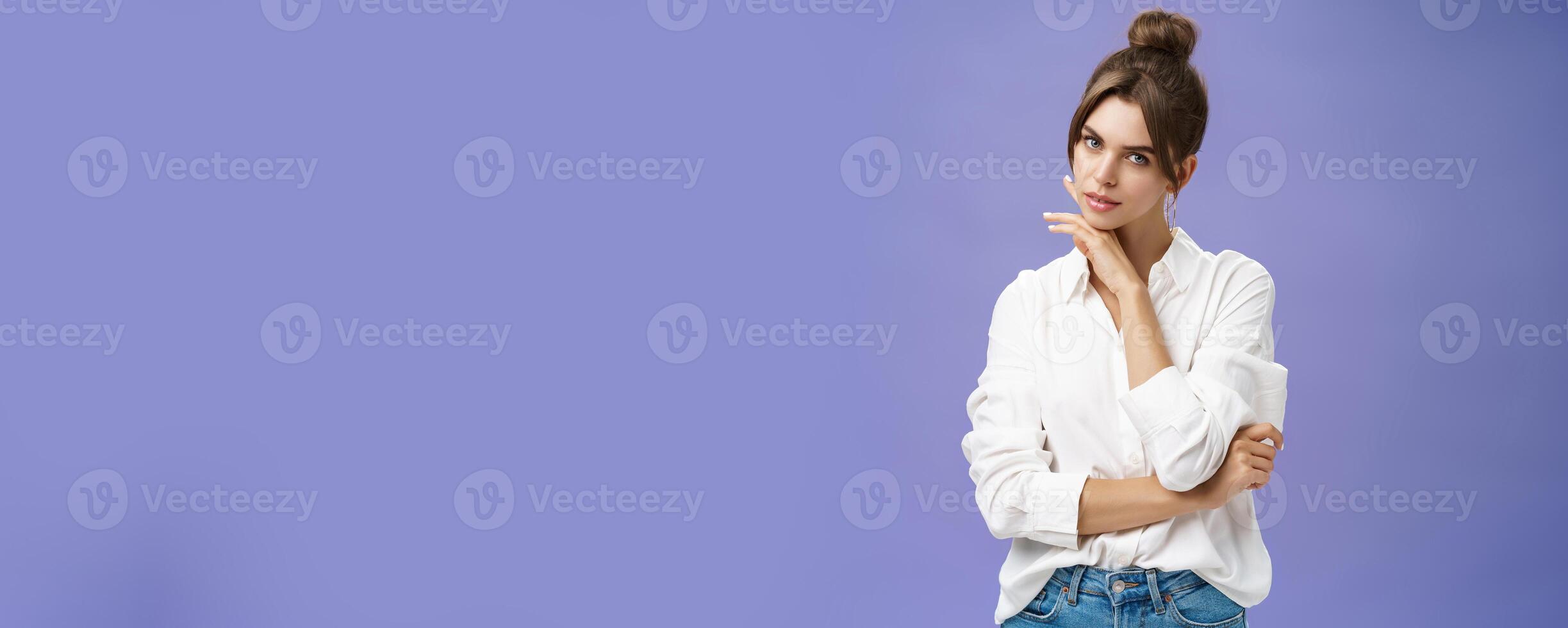 Portrait of tender and feminine stylish woman in white blouse posing sensually and flirty touching chin gazing daring at camera posing against purple background with self-assured expression photo