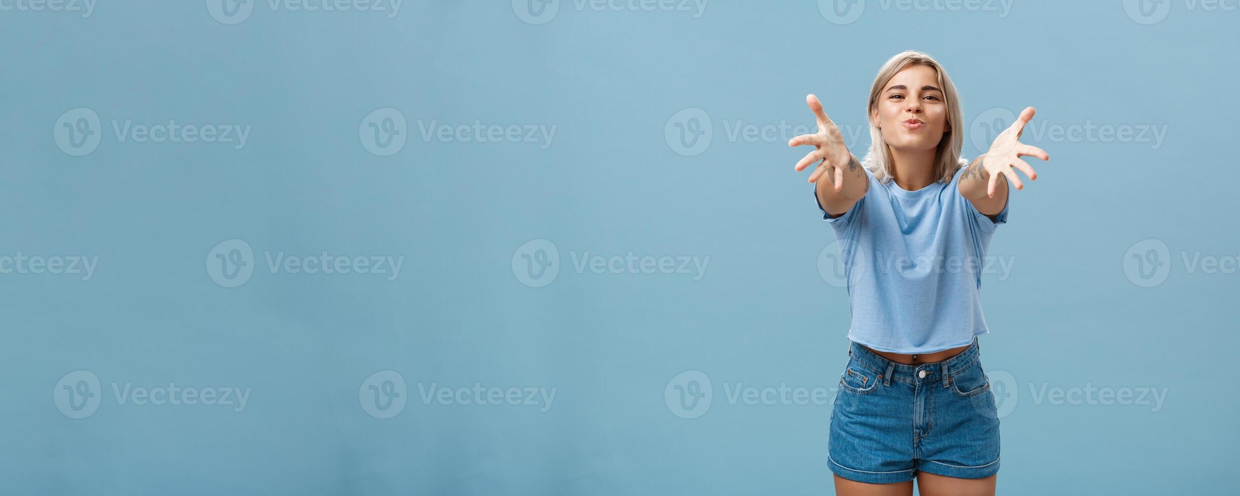 Come to mama. Portrait of friendly joyful and enthusiastic caucasian female student in trendy outfit pulling hands towads camera and folding lips to hug and give passionate kiss smiling over blue wall photo