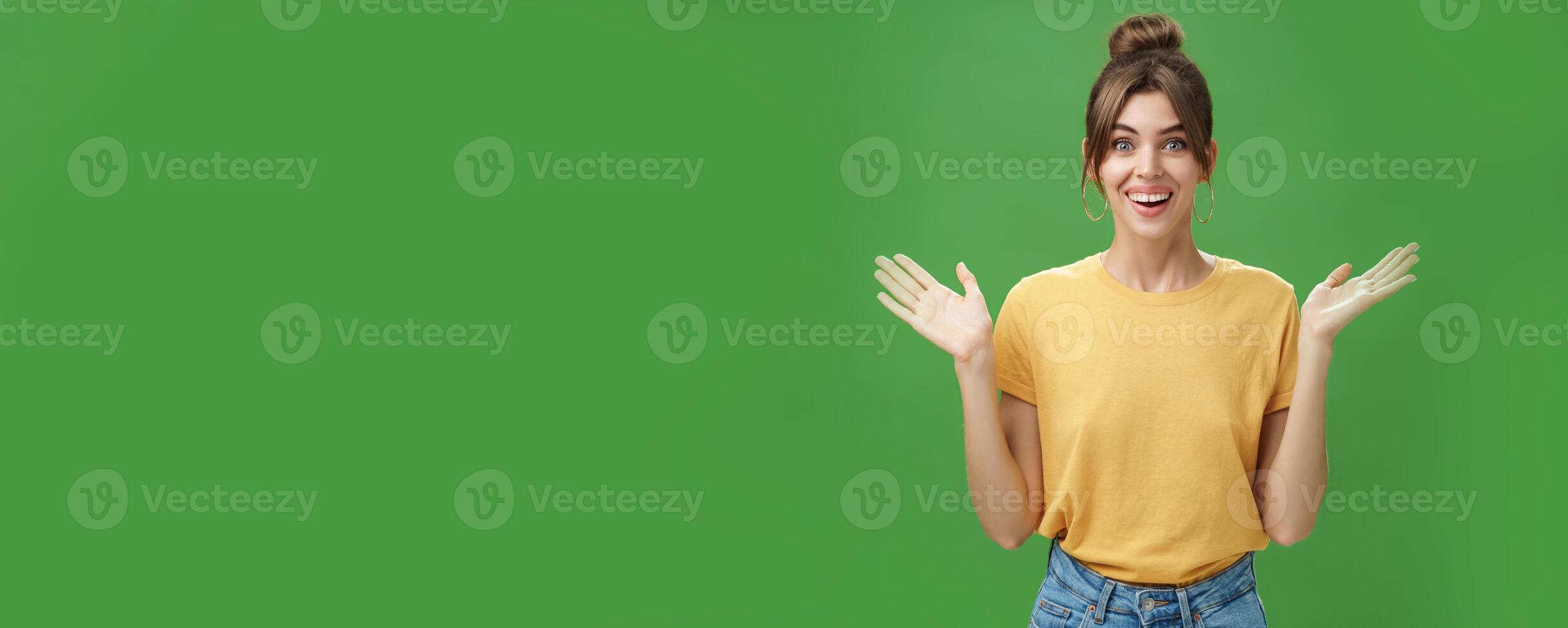 Amazed and surprised woman reacting to astonishing gift raising hands near shoulders smiling happily at camera being delighted and pleased with awesome surprise made by friends over green wall photo