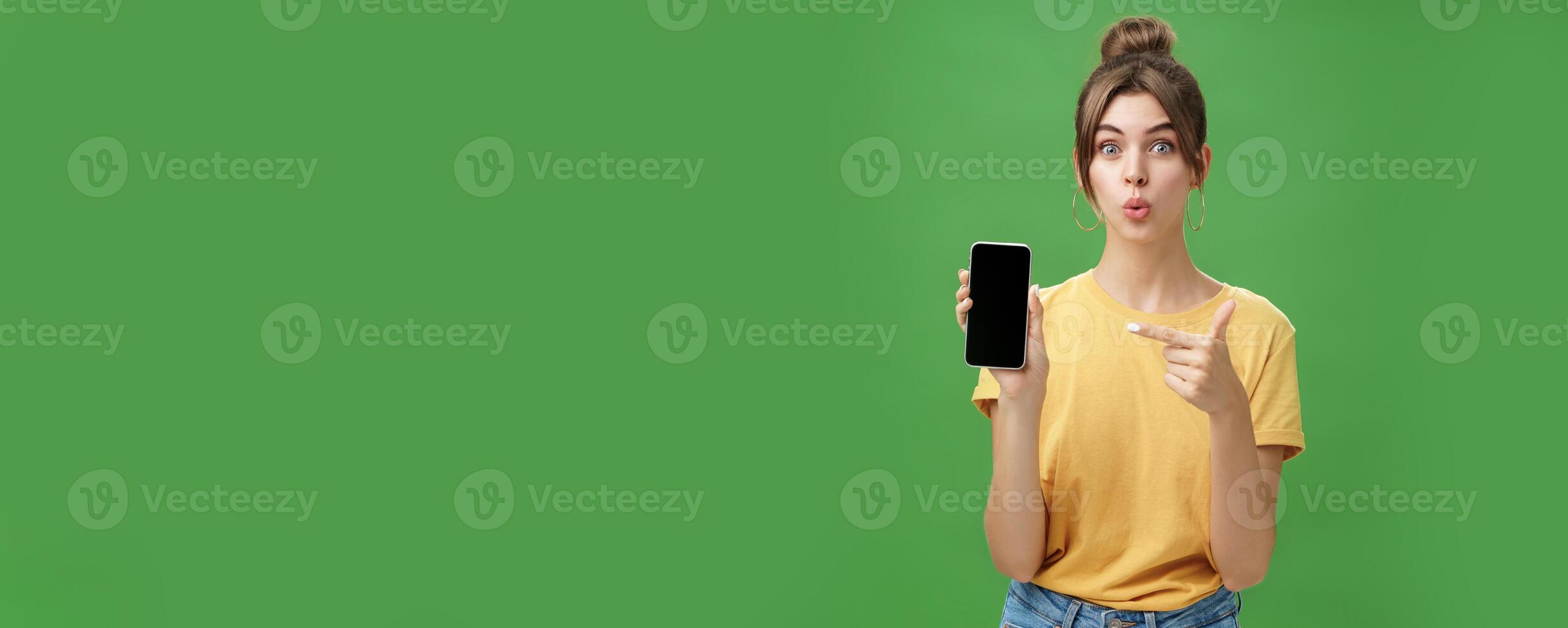 Portrait of intrigued woman cannot wait use new smartphone holding phone and pointing at cellphone screen folding lips with excitement and interest presenting new device model in store over green wall photo