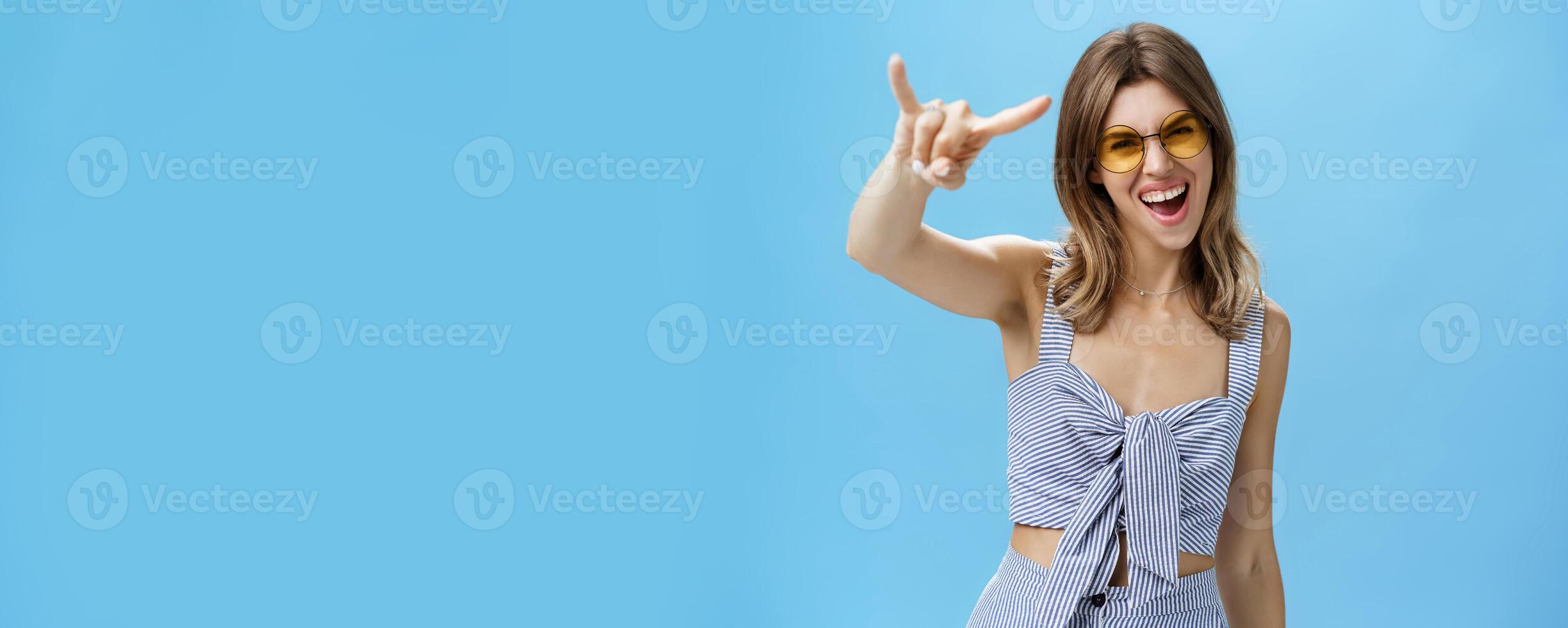 Waist-up shot of confident and daring charming young woman in matching trendy clothes yelling yeah showing rock-n-roll gesture and gazing at camera wearing cool sunglasses enjoying party or festival photo