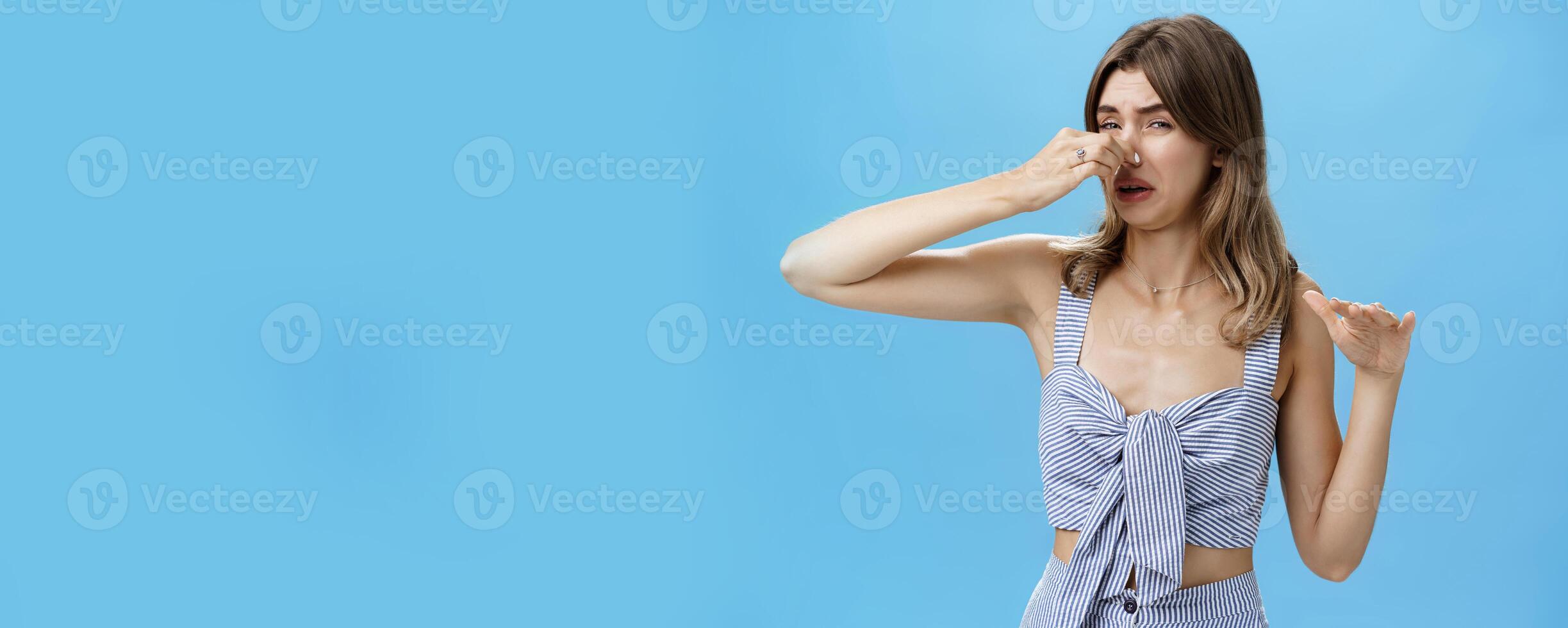Gross smells awful. Portrait of displeased funny young slim woman with tattoo squeezing nose with two fingers not sniff disfusting reek crying from stink standing intense over blue background photo