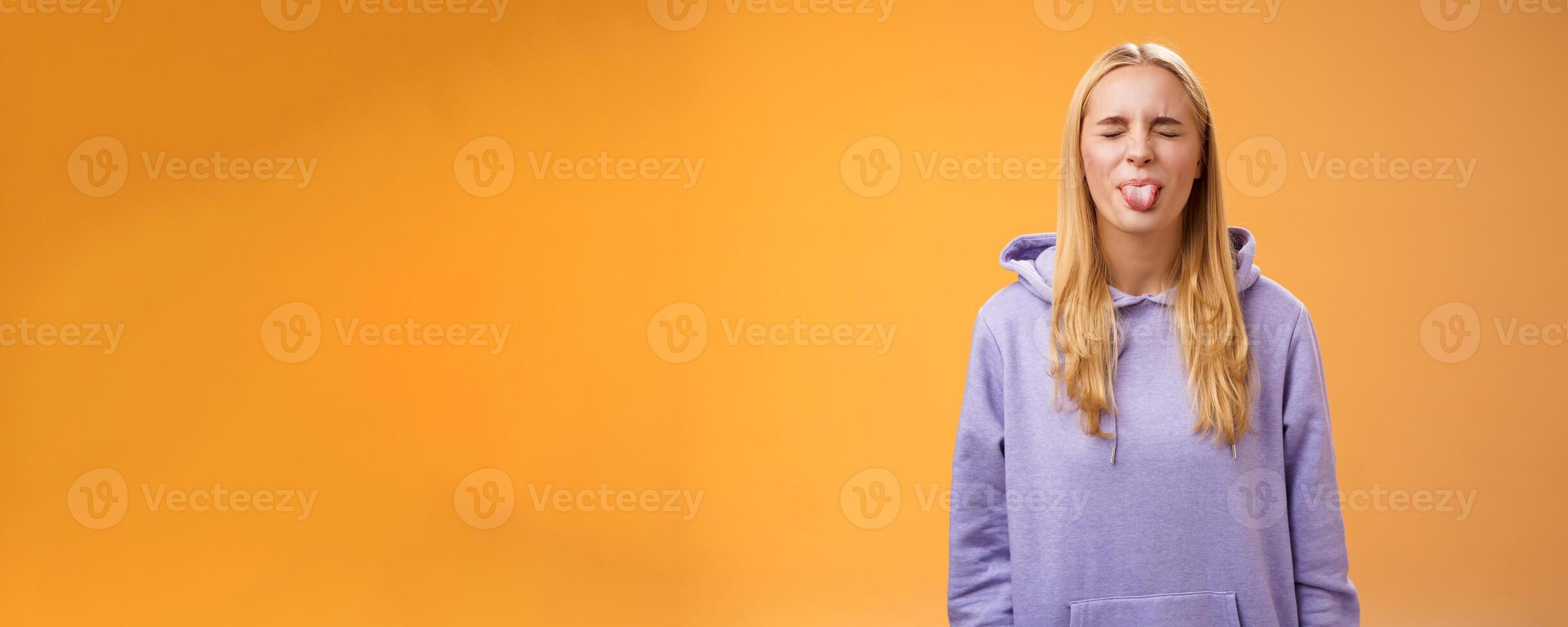 Playful amused charismatic young funny blond girl having fun close eyes showing tongue rebellious fool around unbothered enjoying carefree sunny spring days standing orange background photo