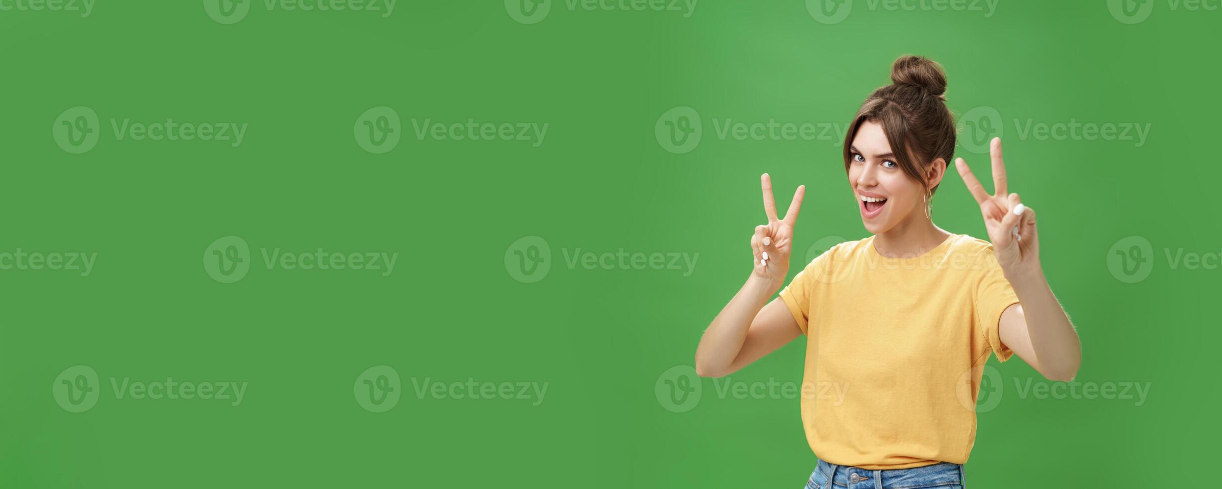 Indoor shot of enthusiastic excited and happy daring girl with combed hair tattoo and cute diasdema showing peace signs bending backwards standing in cool energized pose over green background photo