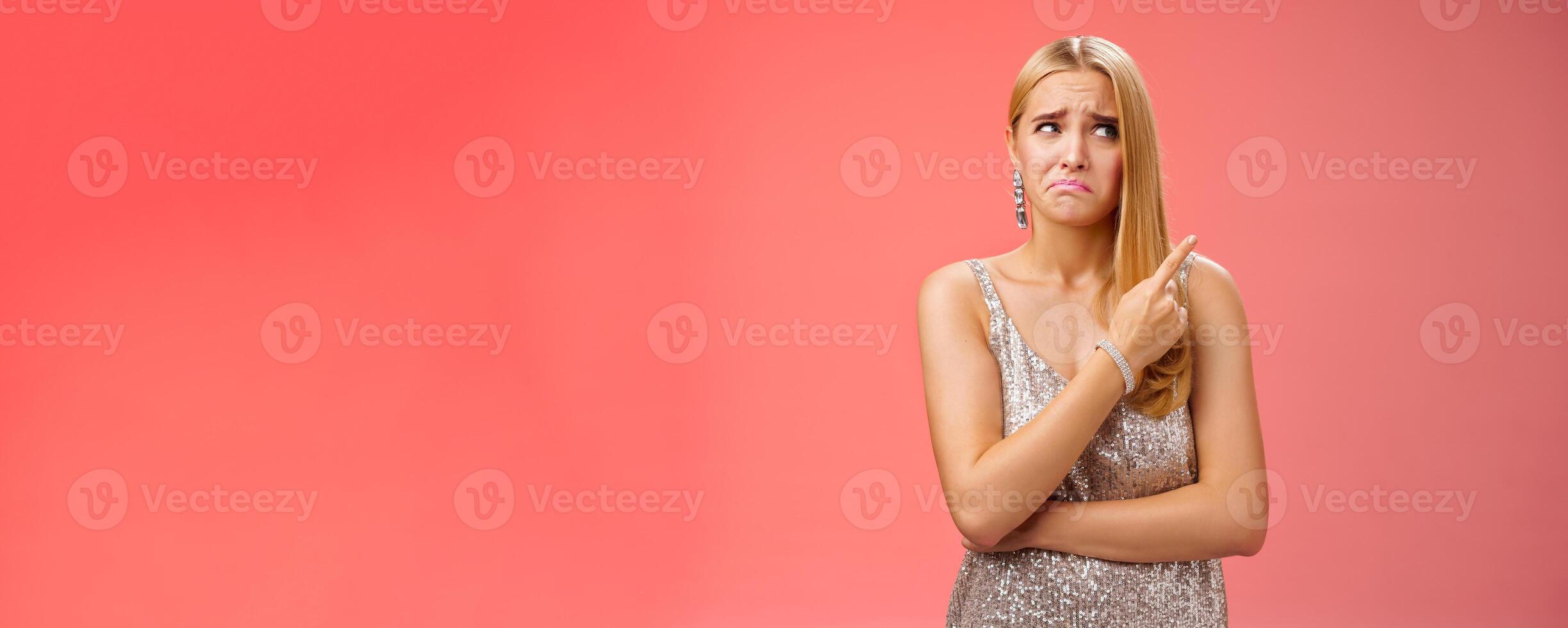 Upset doubtful unsure cute blond funny glamour girl in silver evening dress cringing grimacing hesitant pointing looking upper right corner suspicious, uncertain standing red background photo