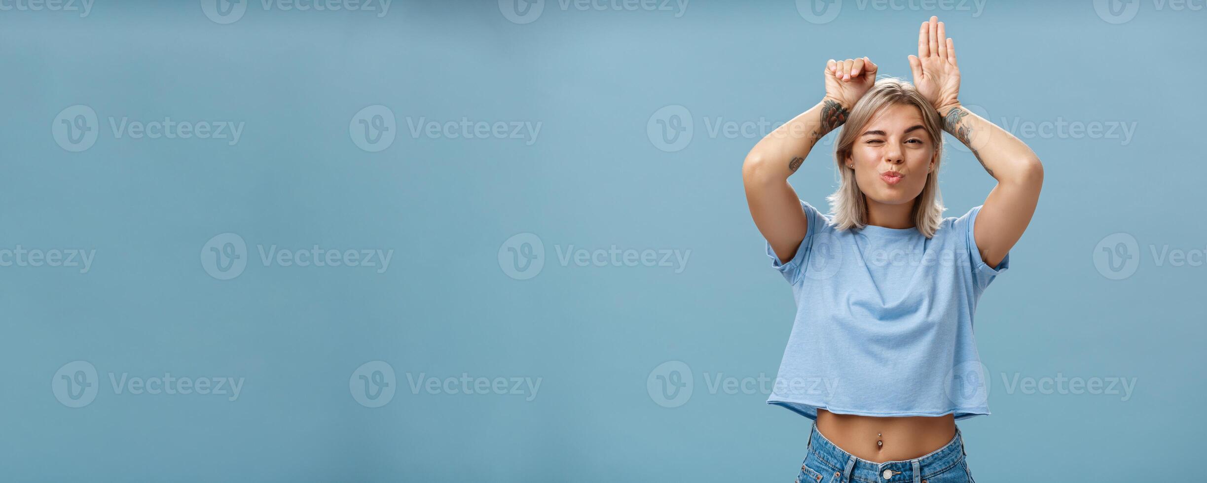 Flirty attractive and stylish cool girlfriend with tattooed arms and pierced belly winking folding lips in air kiss showing bunny ears with palms on head standing over blue background playful photo
