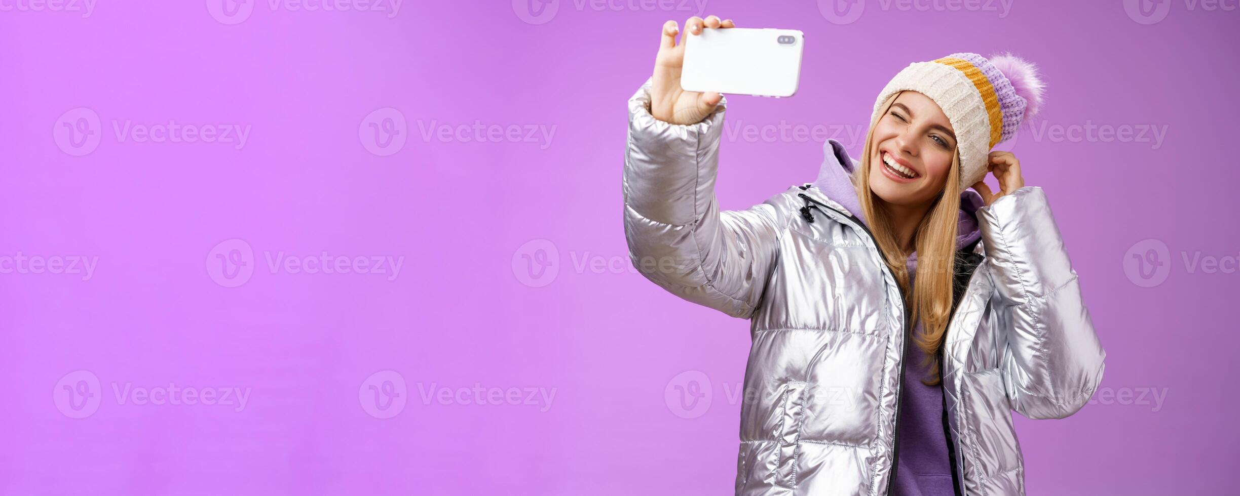 Joyful carefree charming blond girl having fun wanna post pictures from ski resort vacation taking seflie holding smartphone mimicking show tongue winking look mobile display, purple background photo