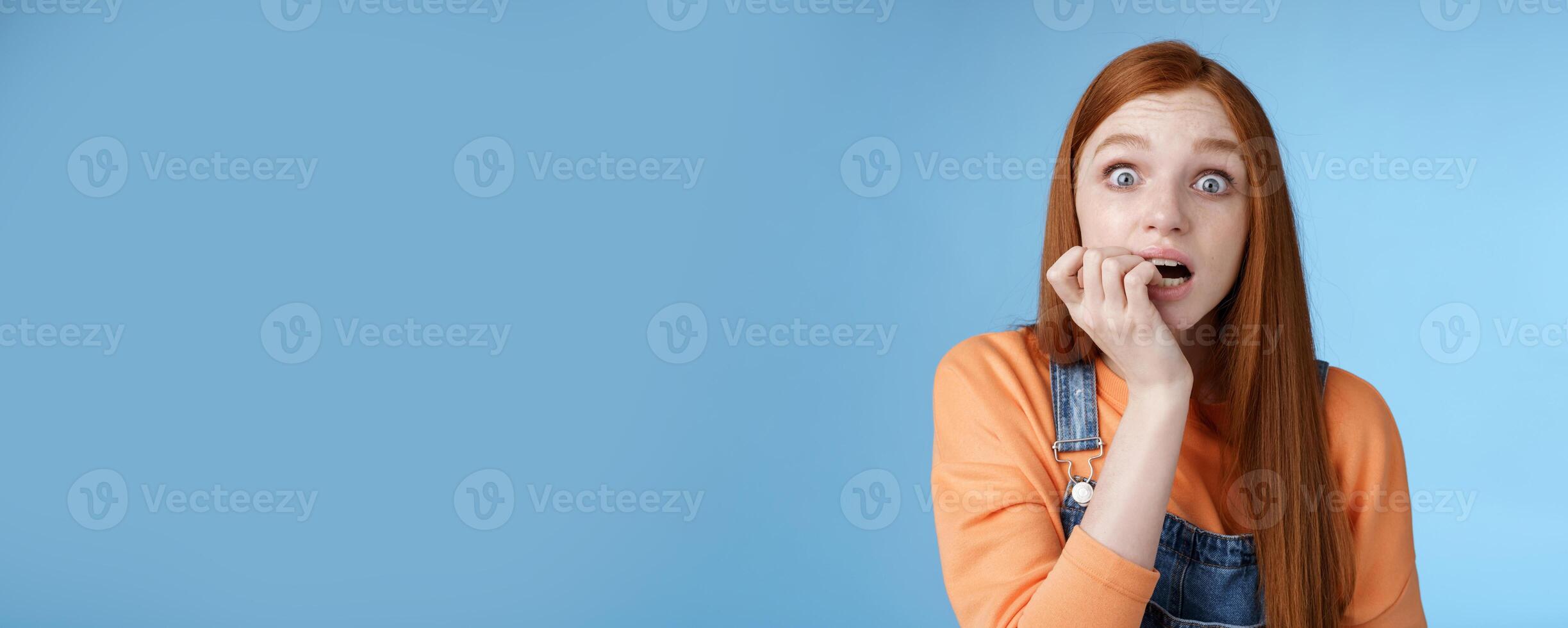 Scared unconfident anxious young trembling redhead girl wide eyes staring intense emotional biting fingernails, fan worry favorite character tv series dies standing nervously blue background photo
